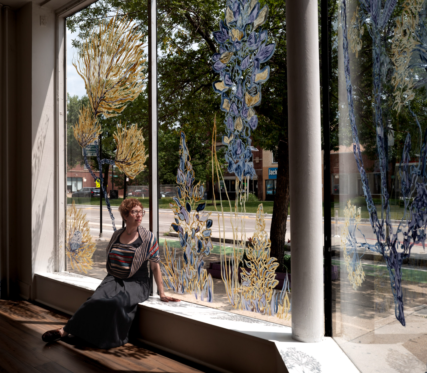 Susy Bielak seated inside Pueblo Unido Gallery beside blue and gold botanical paintings covering the storefront windows of Camas de Ceniza / Ash Beds.