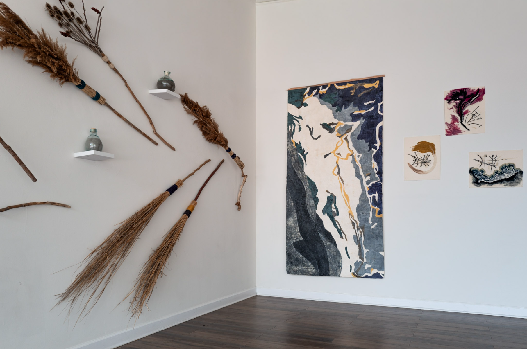 Corner installation view combining a large vertical amate painting, smaller works on paper, handmade broom forms, and bottles of volcanic ash.