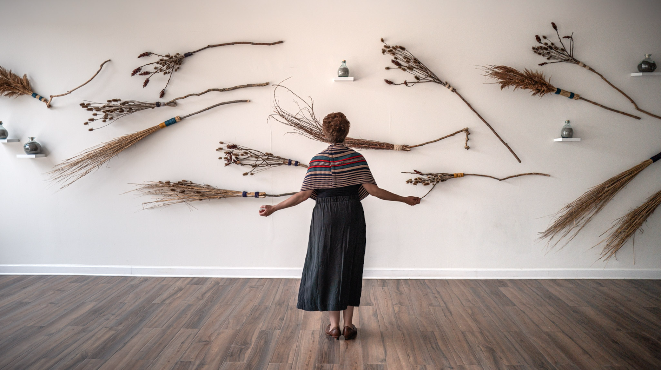 Susy Bielak stands with her back to the camera and arms extended before a wall of handmade broom forms and bottles of volcanic ash.