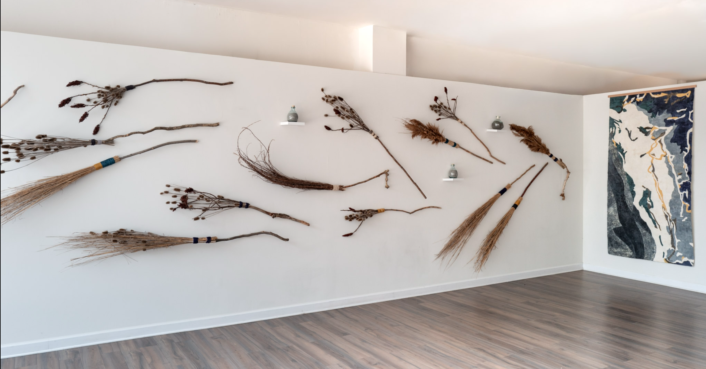 Wide installation view of handmade broom forms and glass bottles of volcanic ash arranged across a white gallery wall, with a large amate painting at right.