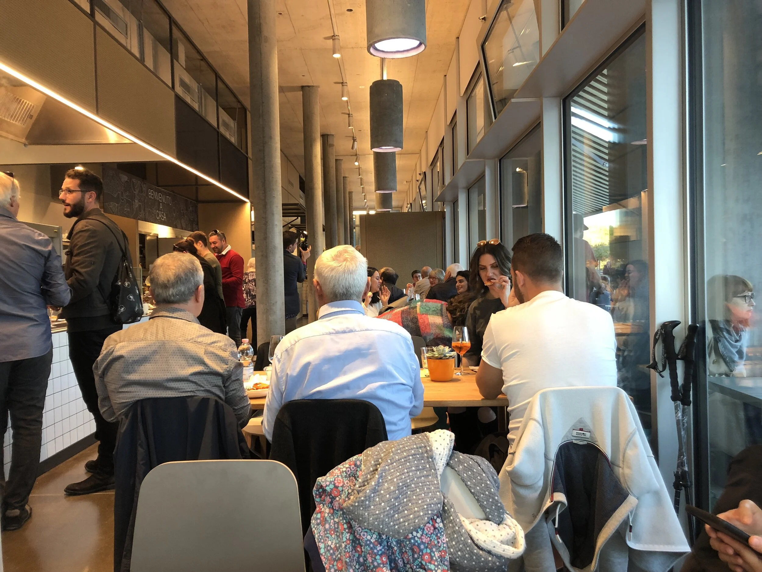 Interior of a busy restaurant with people dining and socializing at tables. The space is filled with natural light from large windows, and there are hanging lights and concrete pillars. A chalkboard sign is visible with writing on it.
