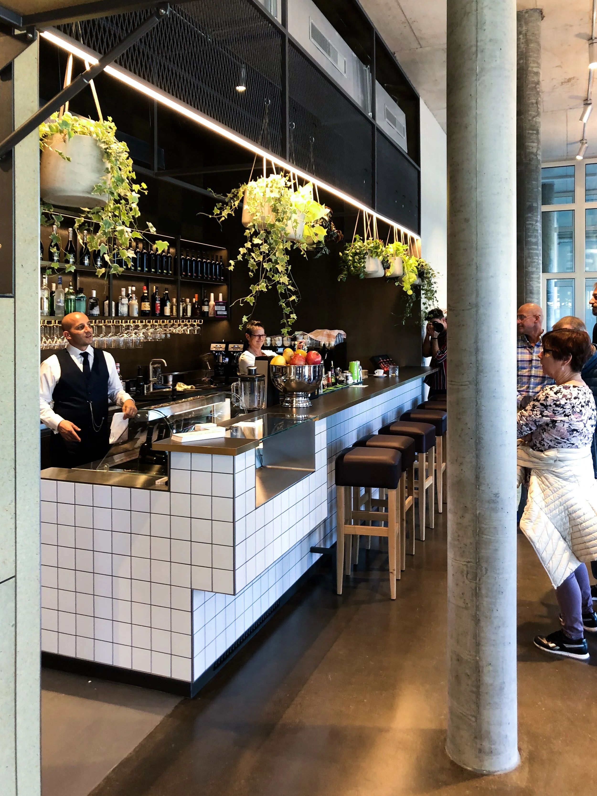 Modern café interior with a tiled bar counter, hanging plants, and seated patrons. A bartender in a vest serves drinks.