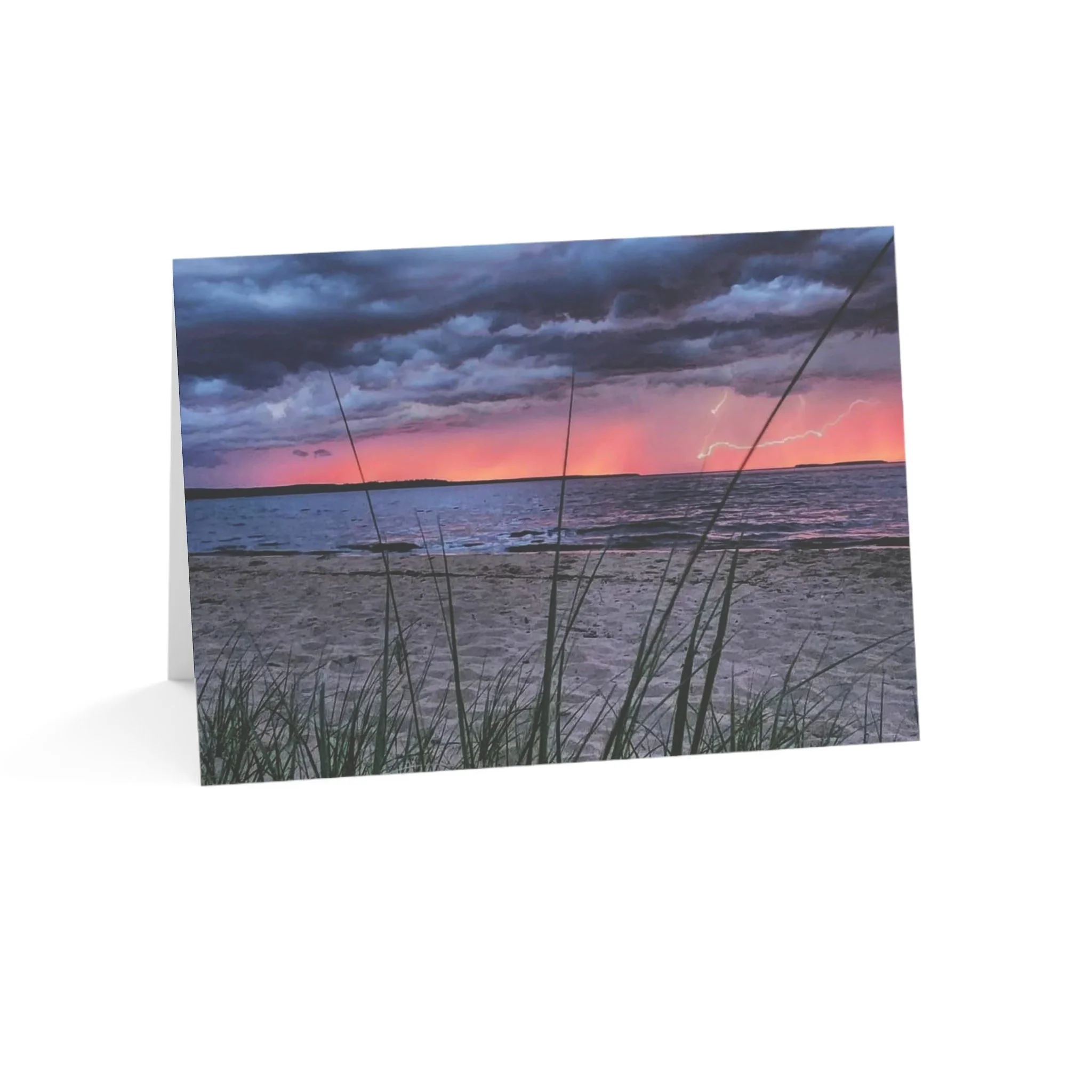 Photo of a beach at sunset with storm clouds, lightning, and pinkish-orange horizon, with grass in the foreground.