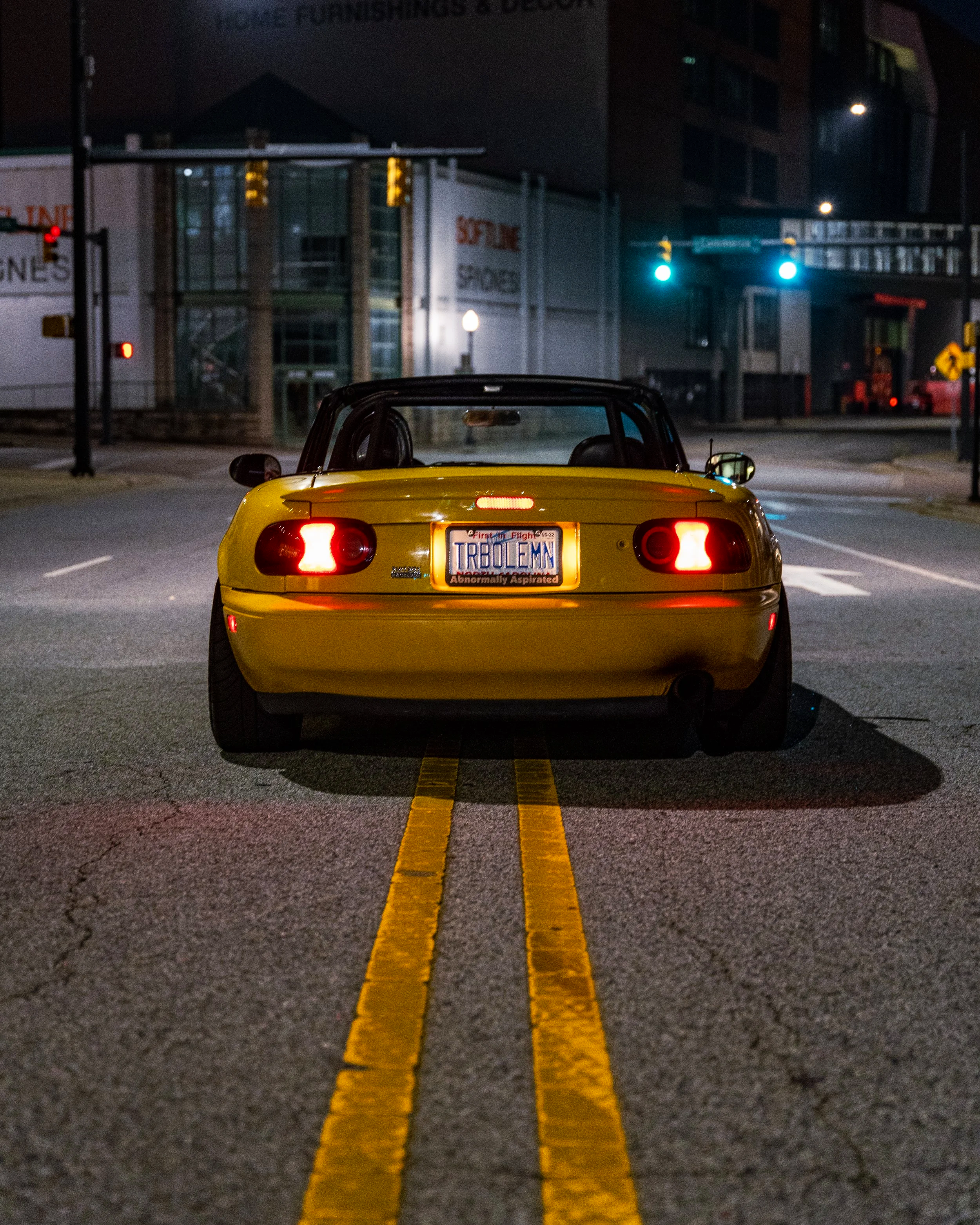 Yellow Mazda Miata parked on a downtown street at night with city lights in the background.