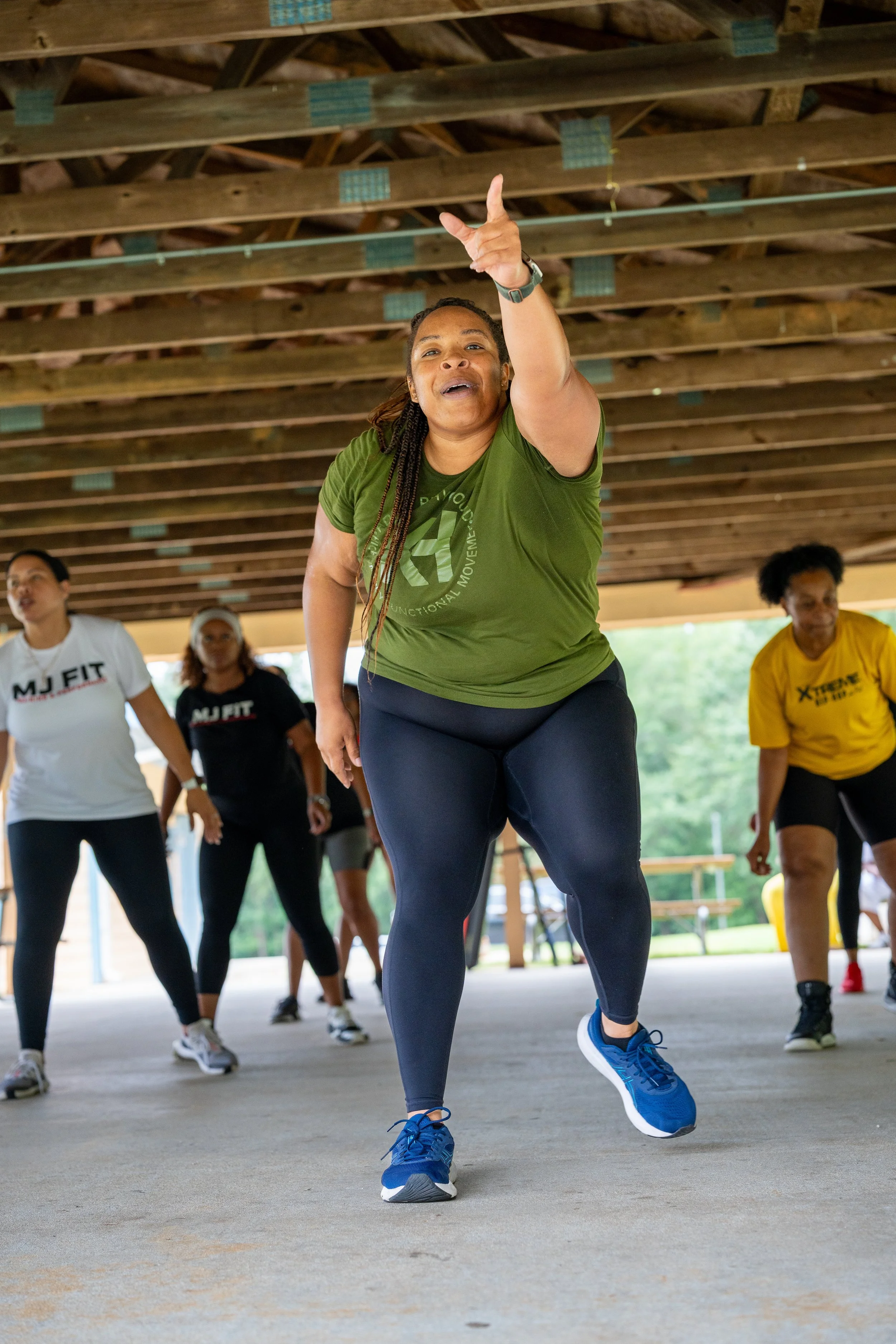 Fitness instructor leading an MJ Fit Training group workout under an outdoor pavilion.