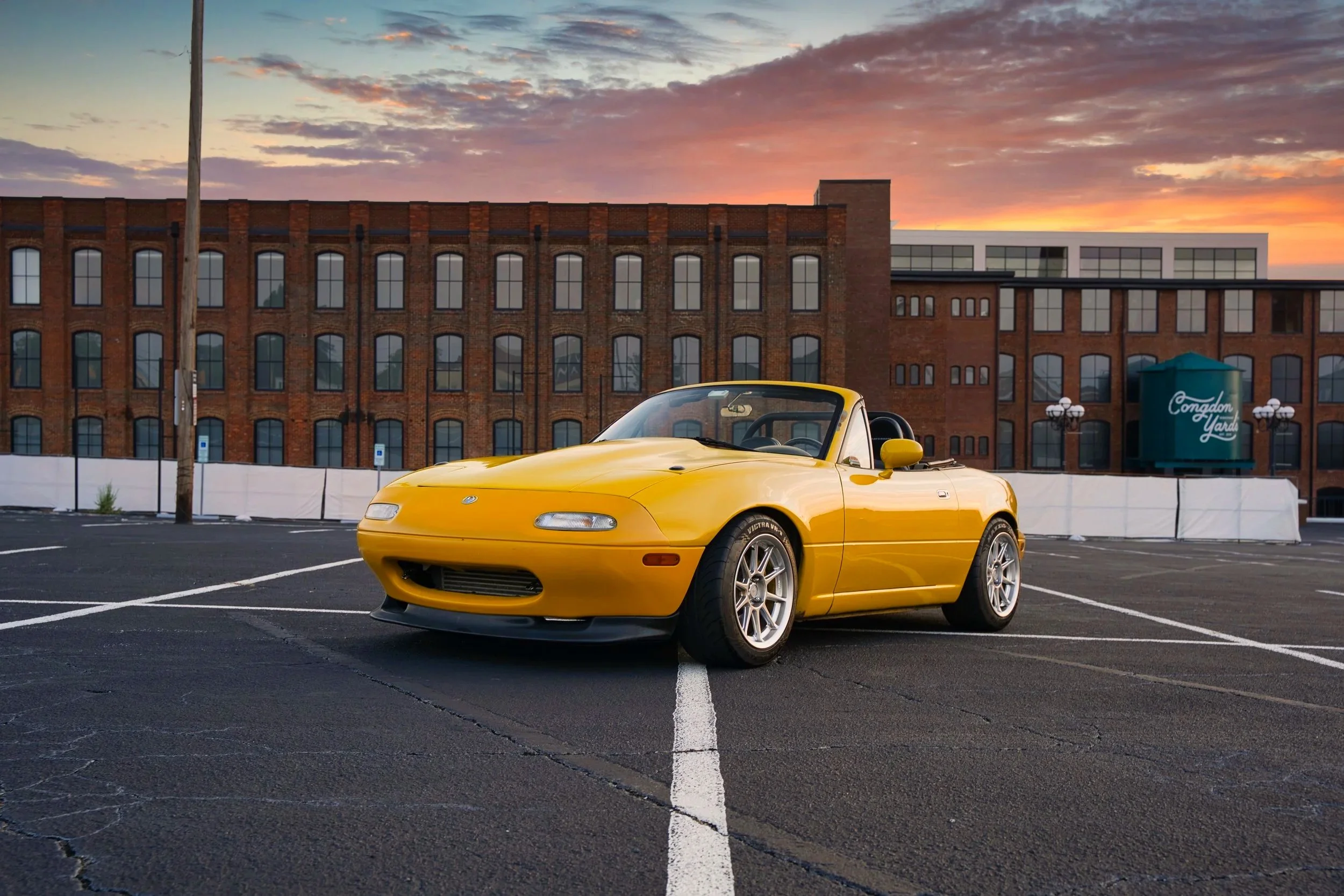 Yellow Mazda Miata photographed in an urban parking lot at sunset.