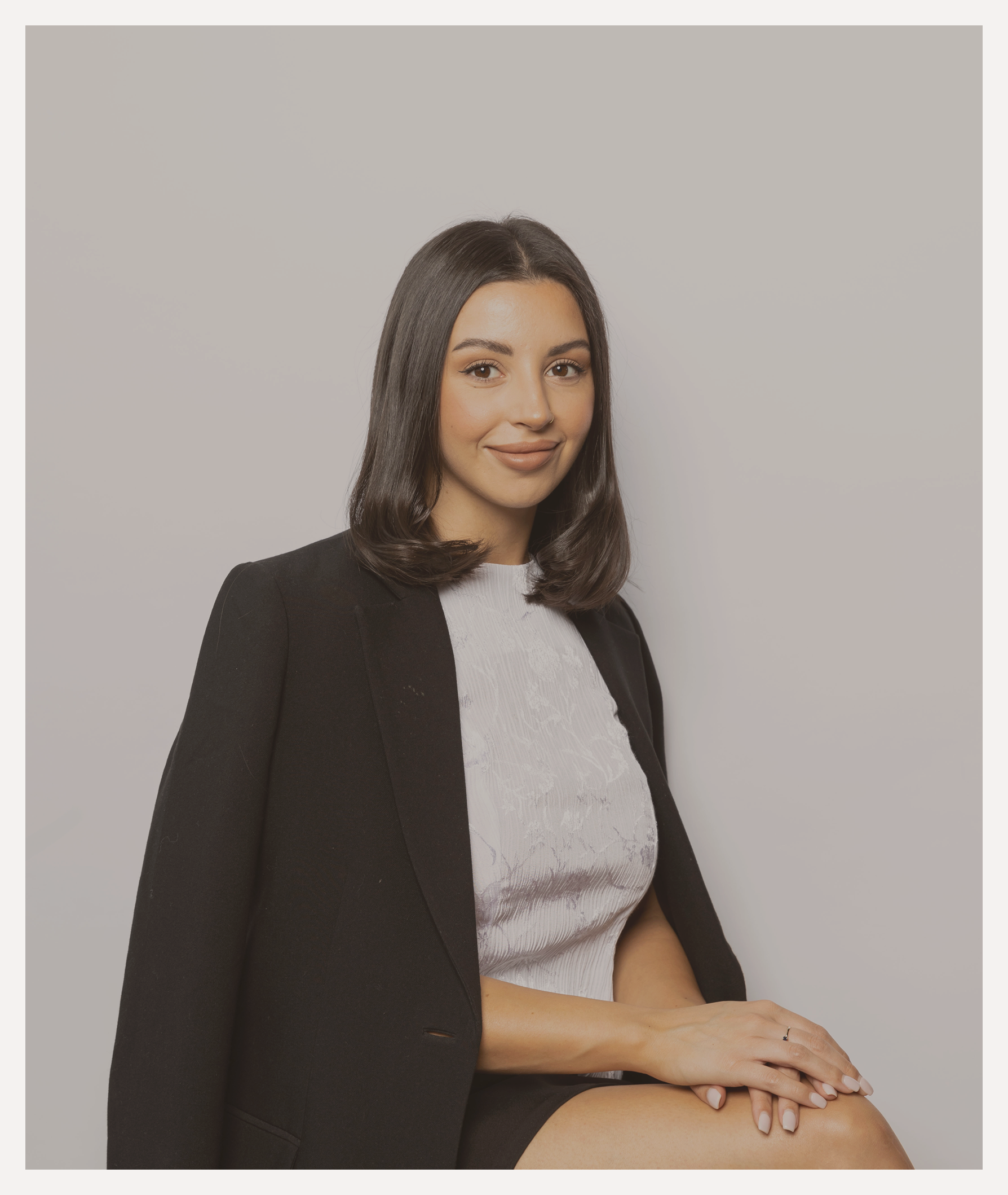 A young woman with shoulder-length dark hair, wearing a black blazer over a light-colored blouse, sitting with one leg crossed over the other against a plain light gray background.