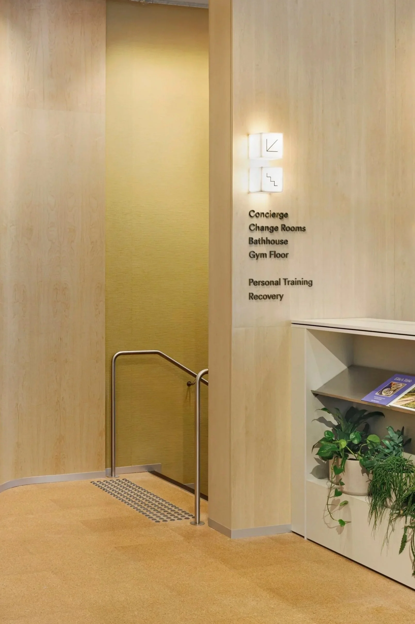 Interior view of a building with a sign listing amenities: concierge, change rooms, bathhouse, gym floor, personal training, and recovery. There is a staircase icon indicating stairs. A small, open reception or information area with plants and brochures is visible on the right.