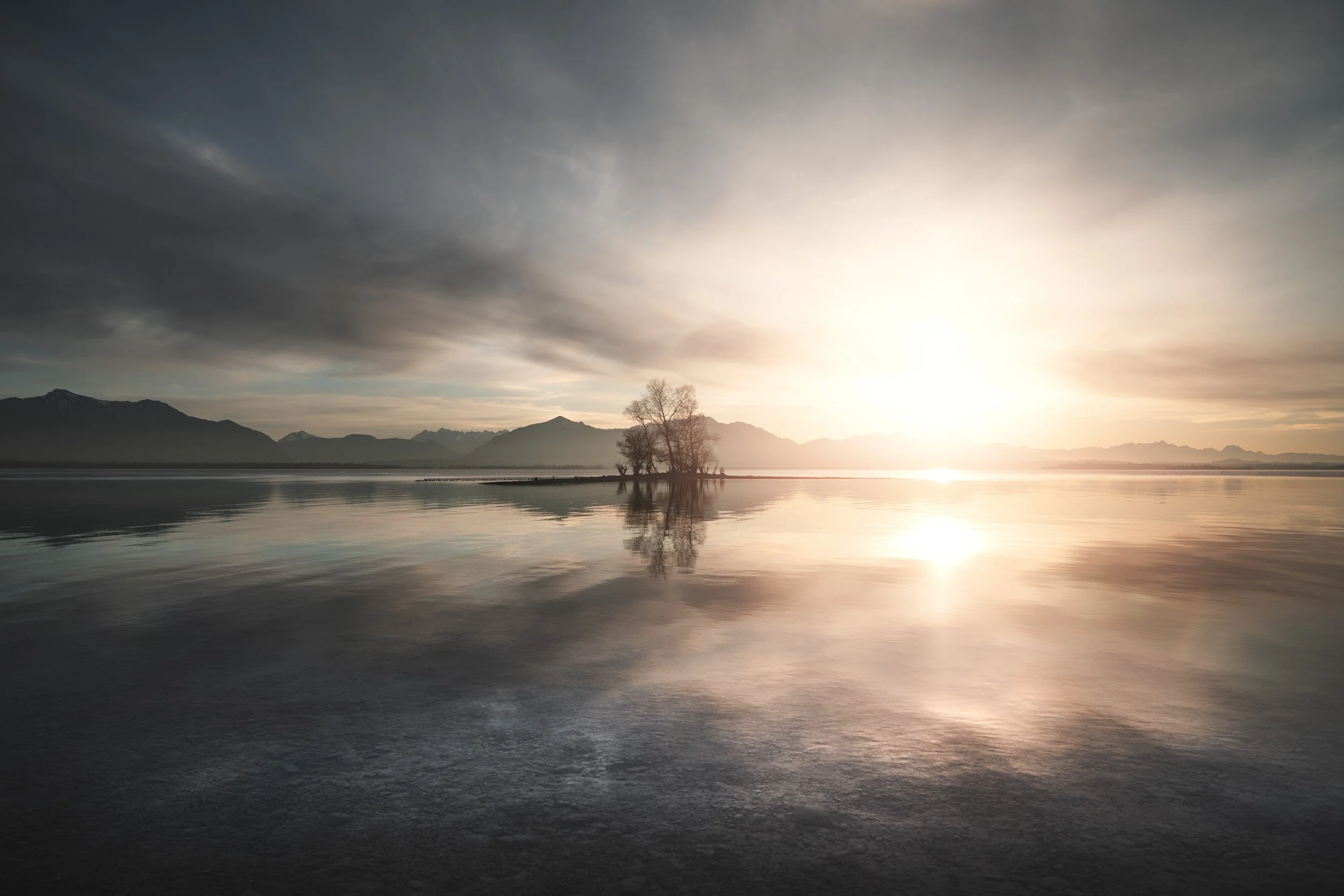 Ein ruhiger See bei Sonnenuntergang mit einem kleinen Baum in der Mitte, umliegende Berge und ein bewölkter Himmel.
