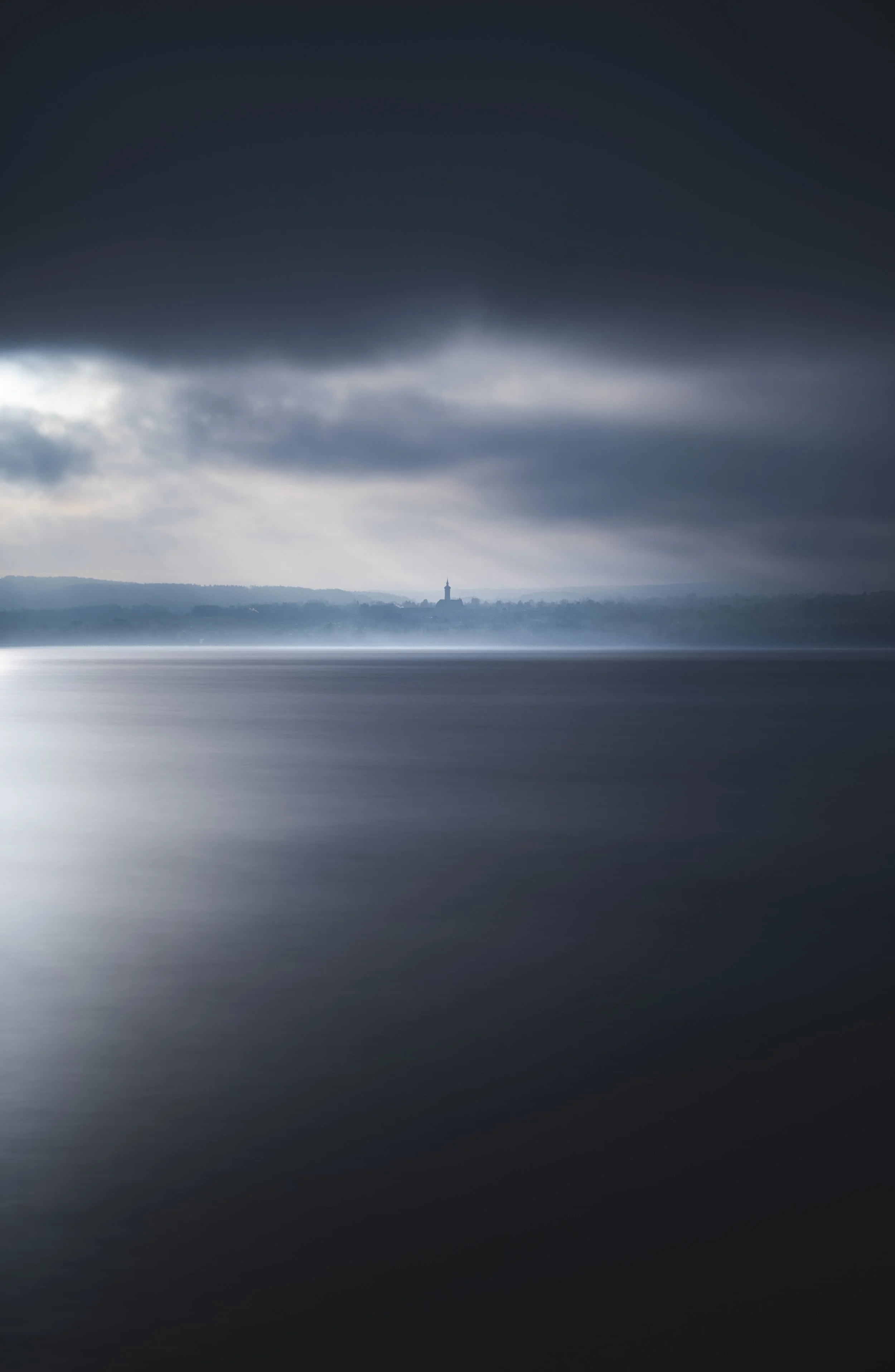 Dunkle Wolken über einem ruhigen Wasser, im Hintergrund eine Kirche am Ufer, stimmungsvoll und melancholisch. Ammersee