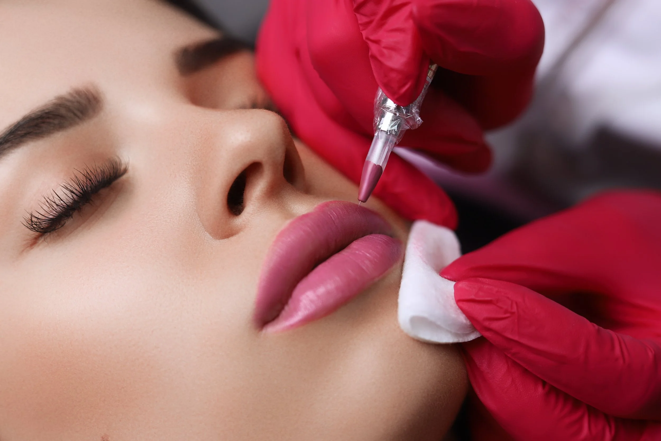 Close-up of a person receiving a permanent lip makeup procedure with a tattoo machine, performed by a professional wearing red gloves.