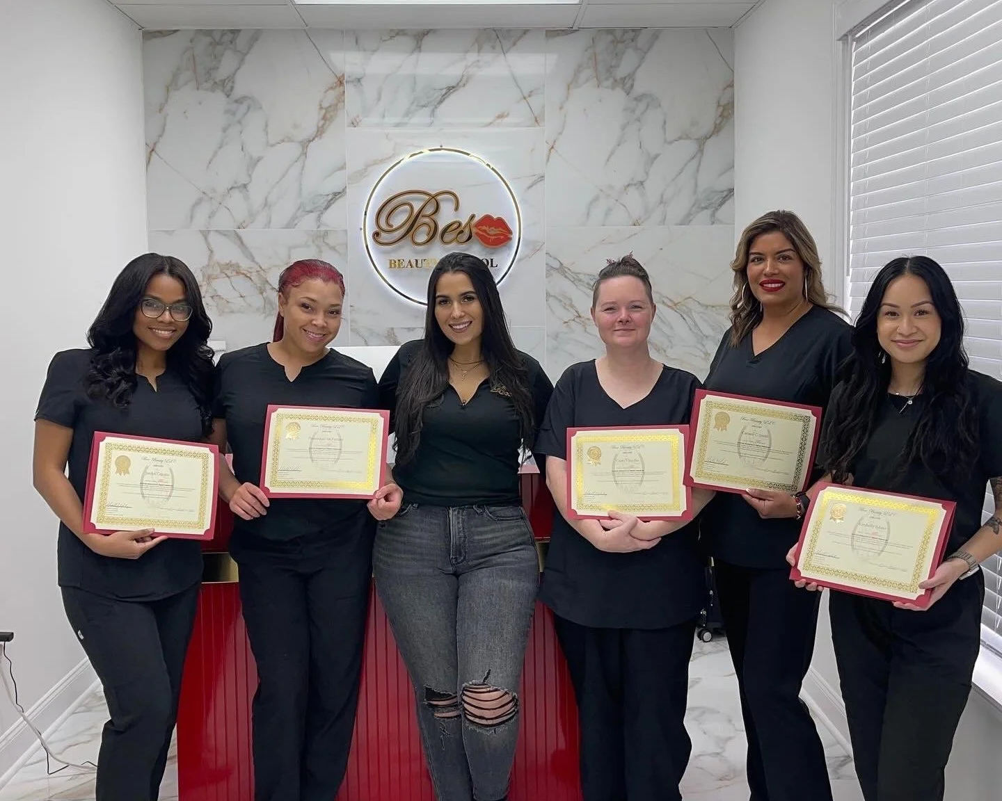 Group of six women holding certificates while wearing black uniforms in a beauty school setting.