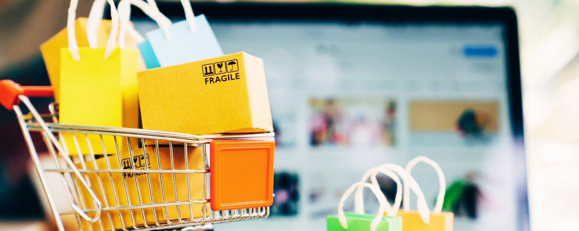 Shopping bags and a small cardboard box in a cart in front of a blurred online retail screen.