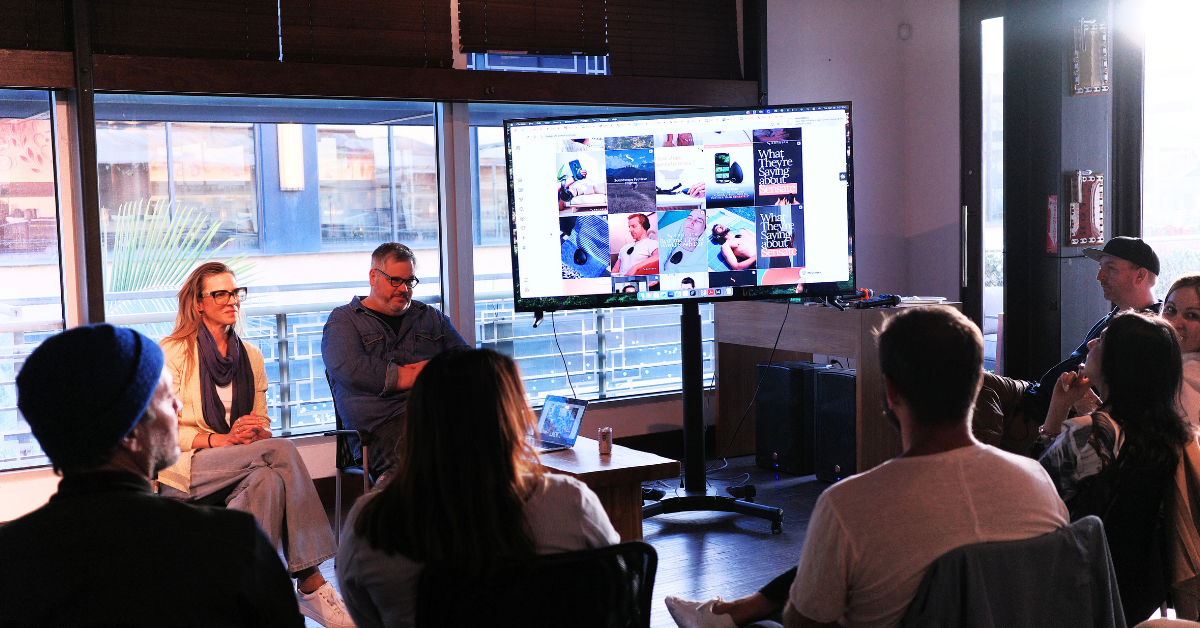 A group of people sit in a conference-style discussion around a large display screen showing a grid of creative visuals and messaging examples.