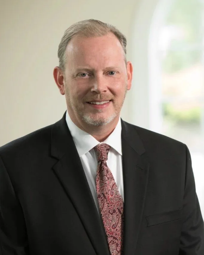 Larry Corthell in a black suit, white shirt, and patterned red tie, smiling confidently in a professional indoor setting with a blurred window in the background.