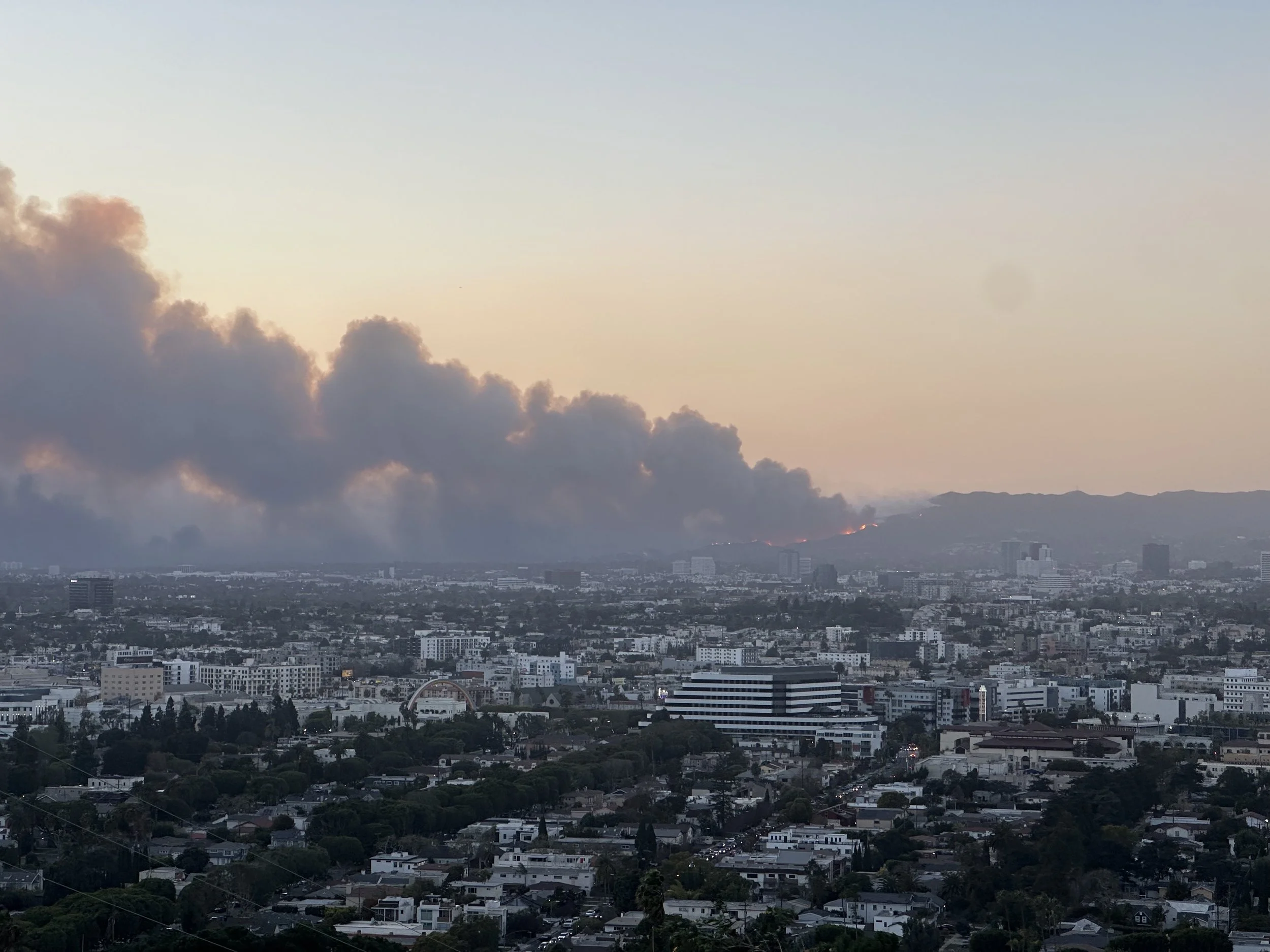 A Los Angeles city skyline at sunset, with a plume of smoke