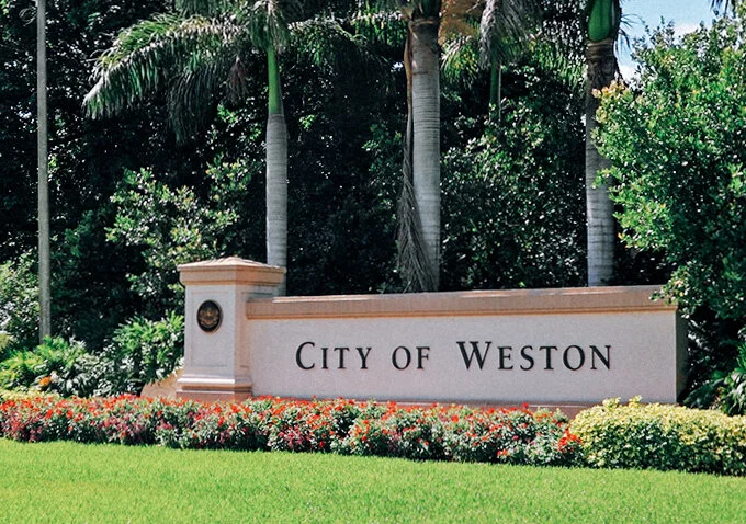 A city sign that reads "City of Weston" is displayed in front of a landscaped area with colorful flowers and tall palm trees in the background.