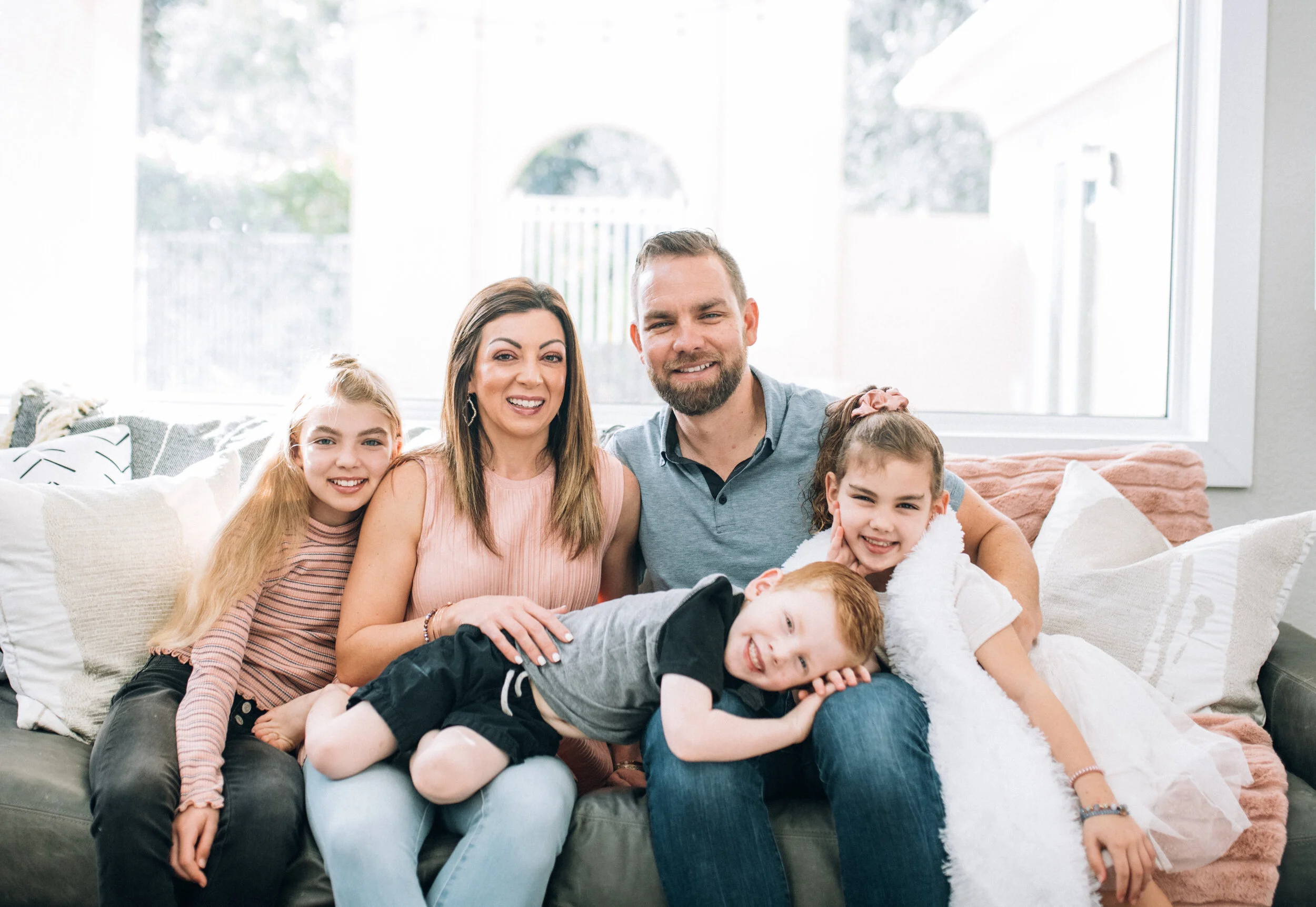 Family of five sitting on a couch in a bright room, smiling at the camera.