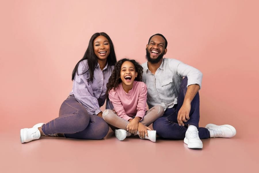 A family of three sitting on the floor and smiling, with a pink background. The mother, father, and daughter are all laughing and appear happy.