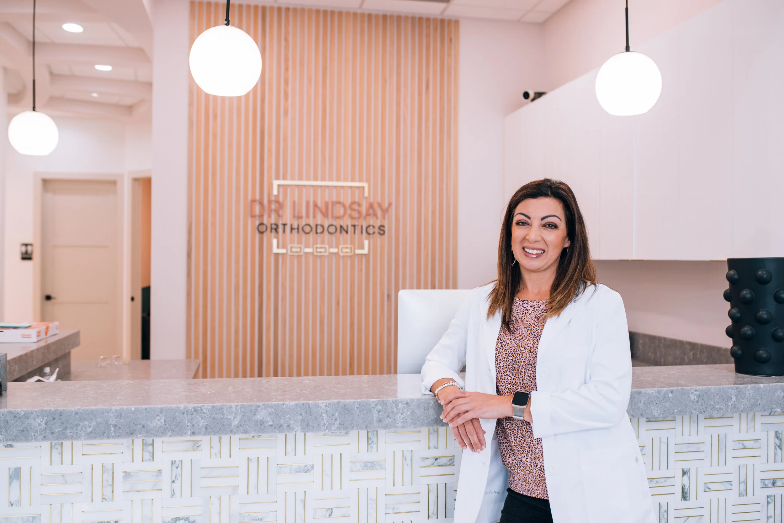 A woman in a white coat standing at a reception desk in a modern orthodontics office, smiling and looking at the camera.