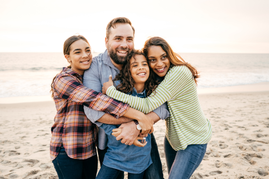 A family of four, consisting of two women, one man, and one girl, smiling and embracing on a sandy beach with the ocean in the background at sunset.