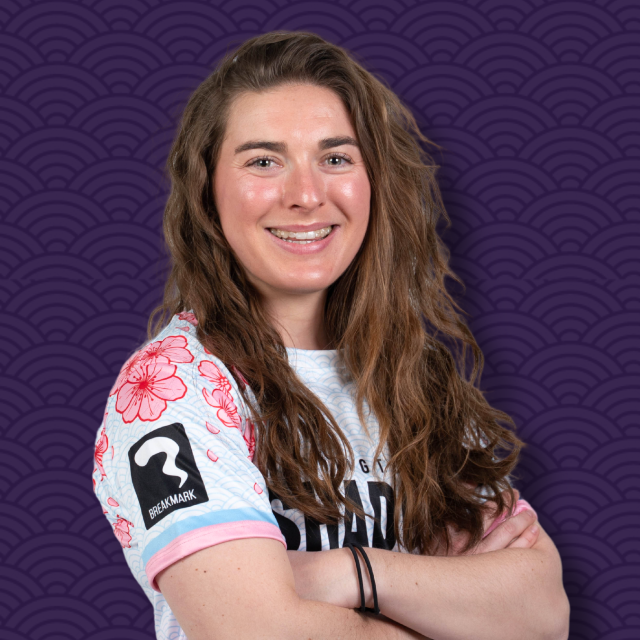 A young woman with long, wavy brown hair smiling and crossing her arms, wearing a colorful, patterned T-shirt against a purple background with a wave pattern.