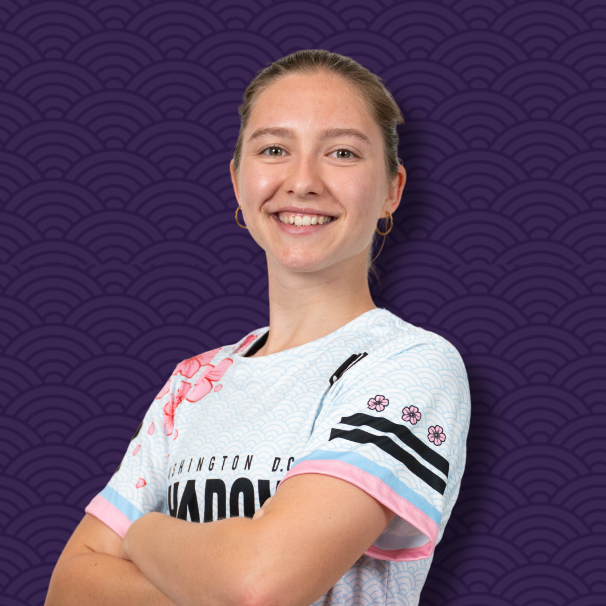 Close-up of a young woman with brown hair, smiling, wearing a colorful t-shirt with floral and Washington DC text, crossing her arms against a purple patterned background.