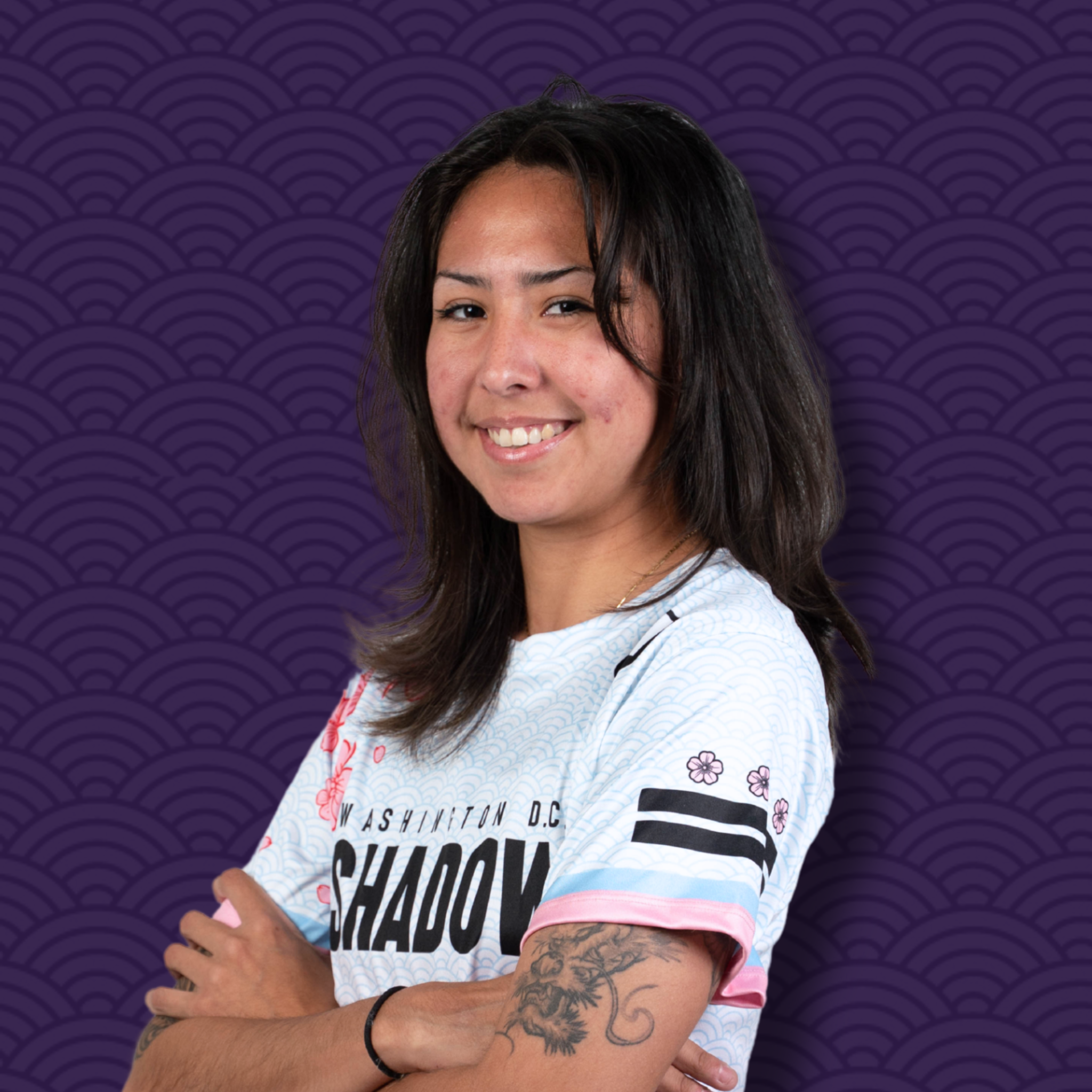 Portrait of a young woman with dark hair, smiling, wearing a white shirt with black and pink text, standing against a purple patterned background.
