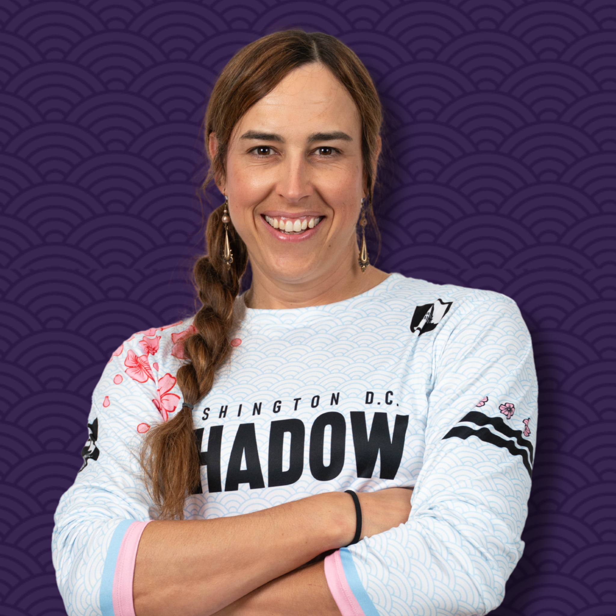 Portrait of a woman with brown hair styled in a braid, smiling, wearing a sports jersey with a sea wave pattern, against a purple patterned background.