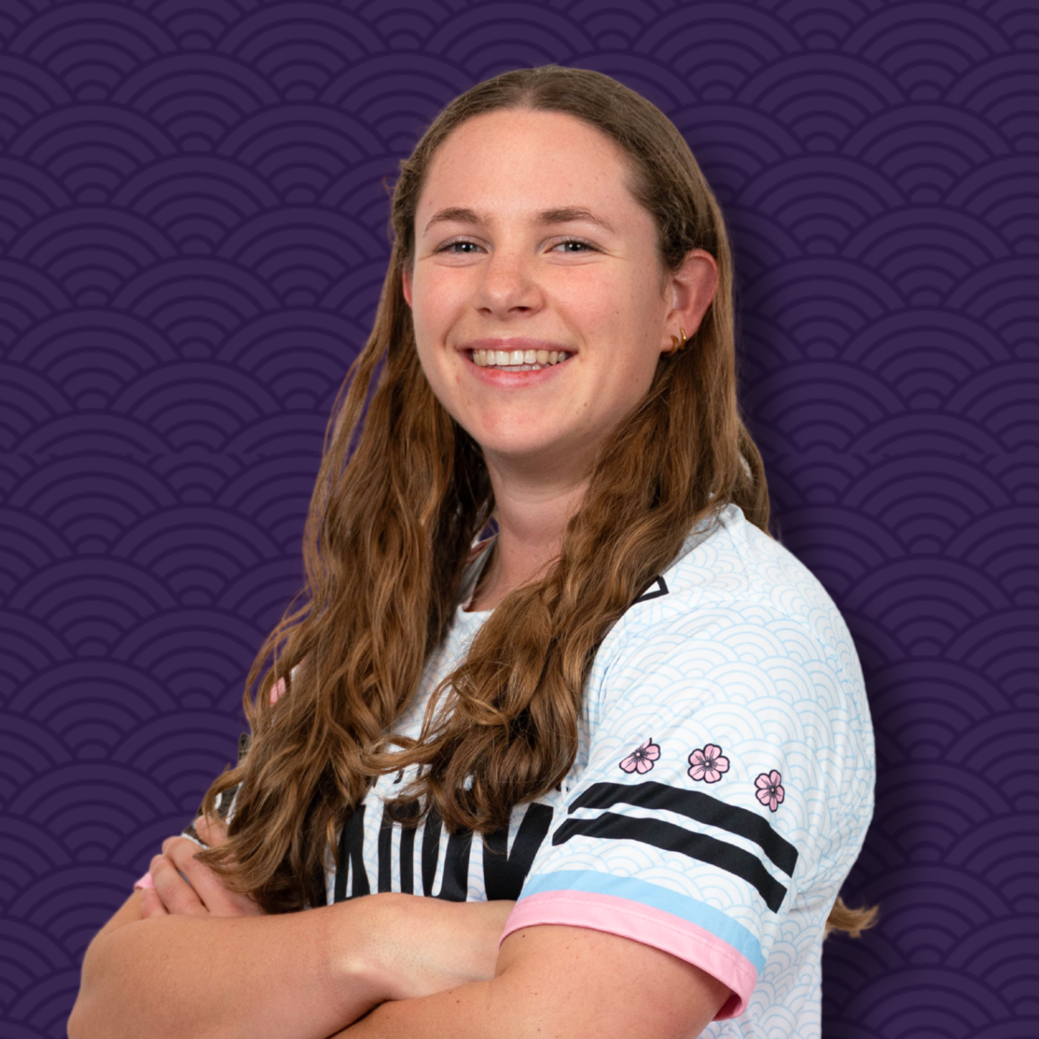A smiling young woman with long wavy brown hair, wearing a white shirt with a light blue pattern, standing in front of a purple background with a repeating scale pattern.