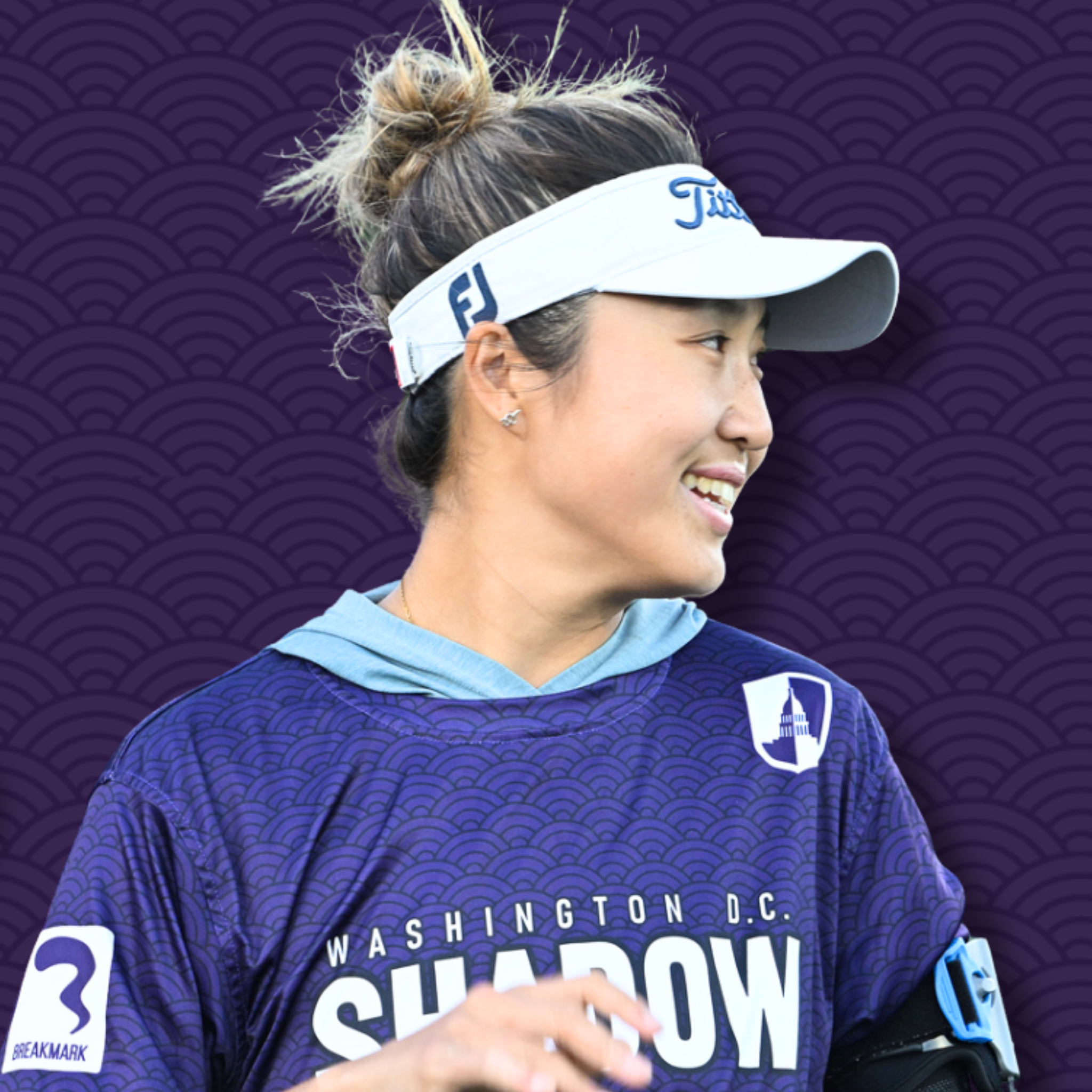 A person wearing a purple jacket, a floral shirt, black gloves, a white visor with the brand 'Titleist', and holding a golf club, standing against a purple patterned background.