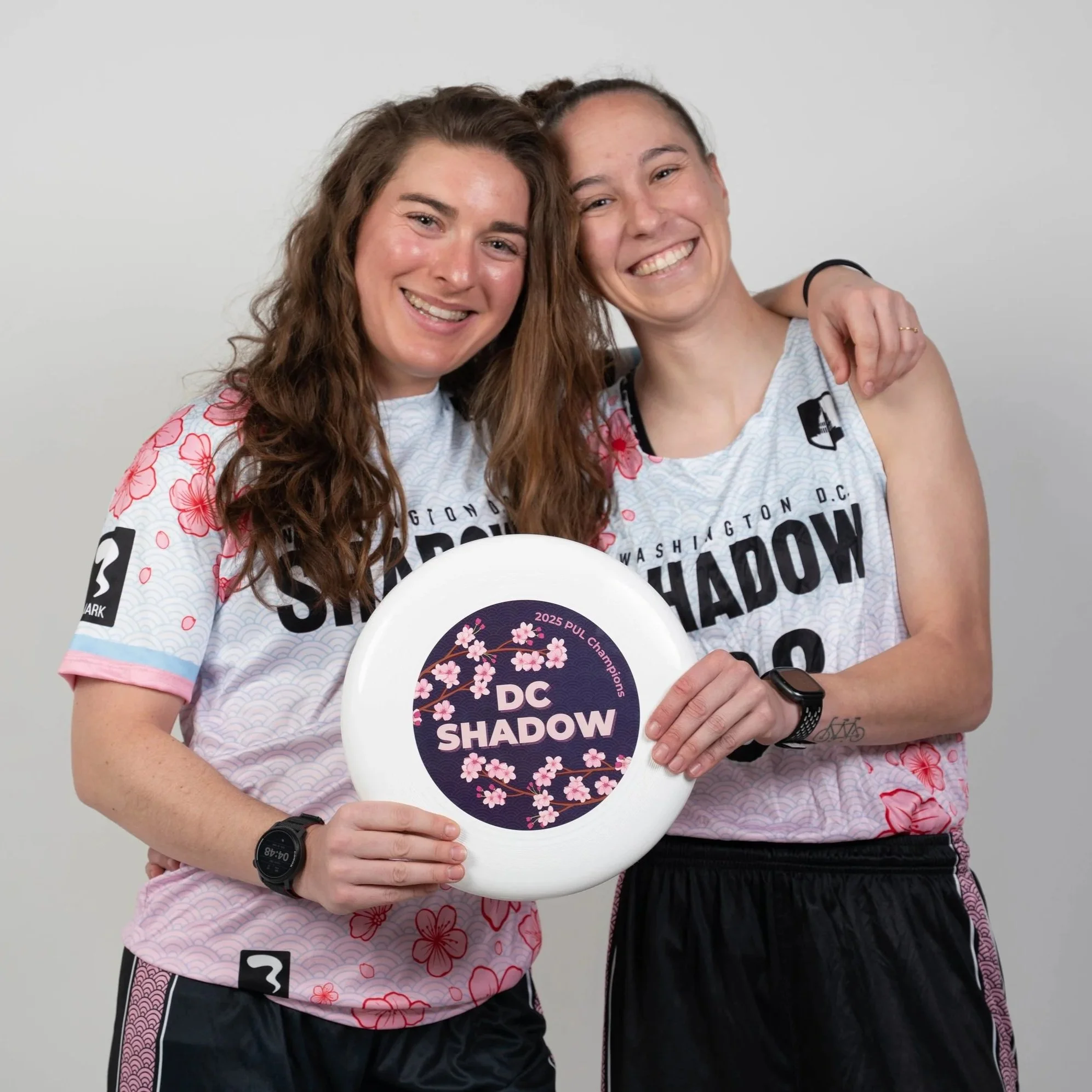 Two women smiling, one with long wavy hair wearing a white and pink floral sports jersey, and the other with hair in a bun wearing a similar sports jersey, are posing together against a light gray background. The woman on the right is holding a disc that reads "2025 Pul Championships" with cherry blossom illustrations, while the woman on the left has her arm around her friend's shoulder.