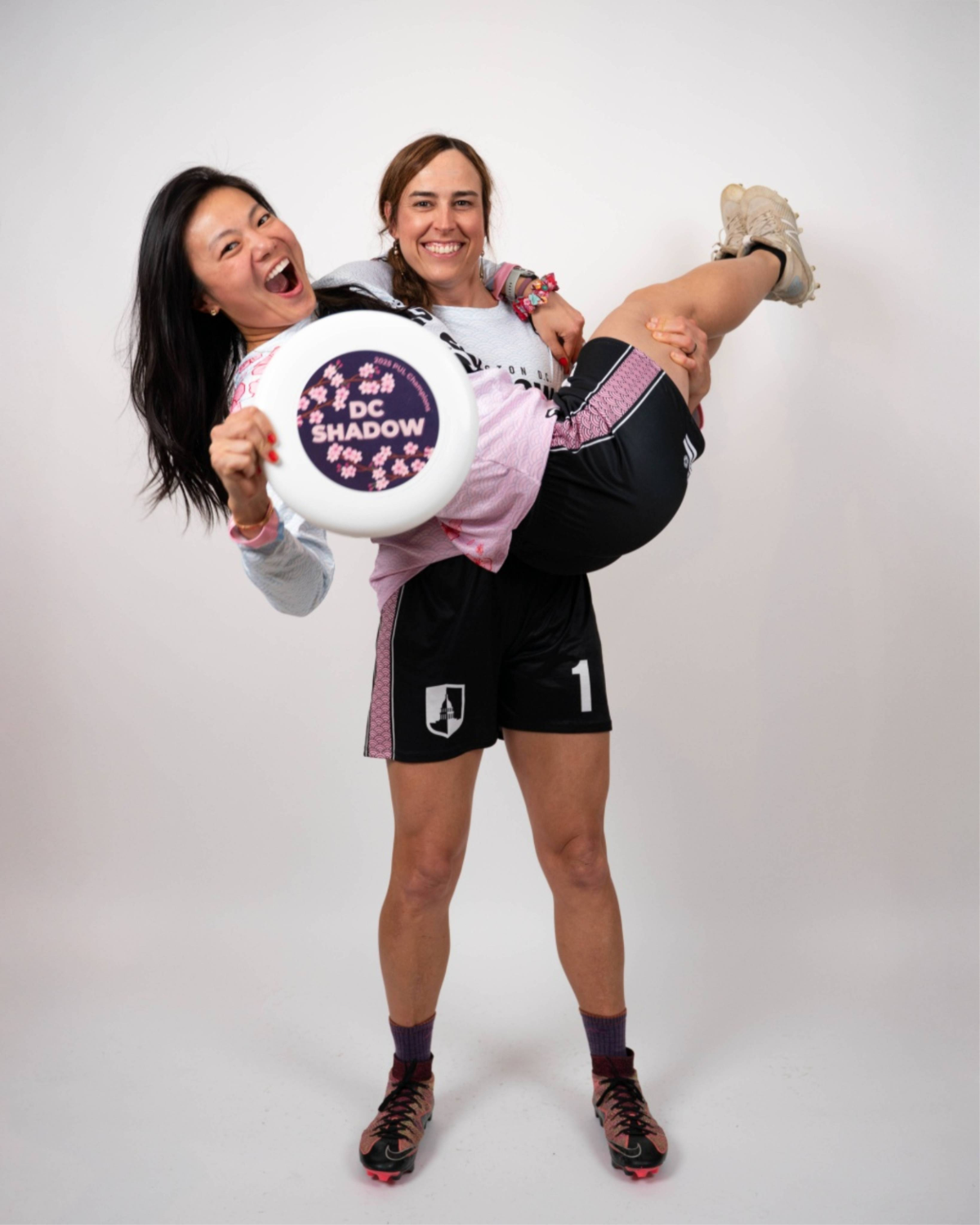 Two women celebrating, one is holding the other in a playful lift. The woman being lifted is holding a frisbee that says "DC SHADOW" with cherry blossom graphics. Both are smiling and dressed in athletic gear, with running shoes and sports jerseys, standing against a plain white background.