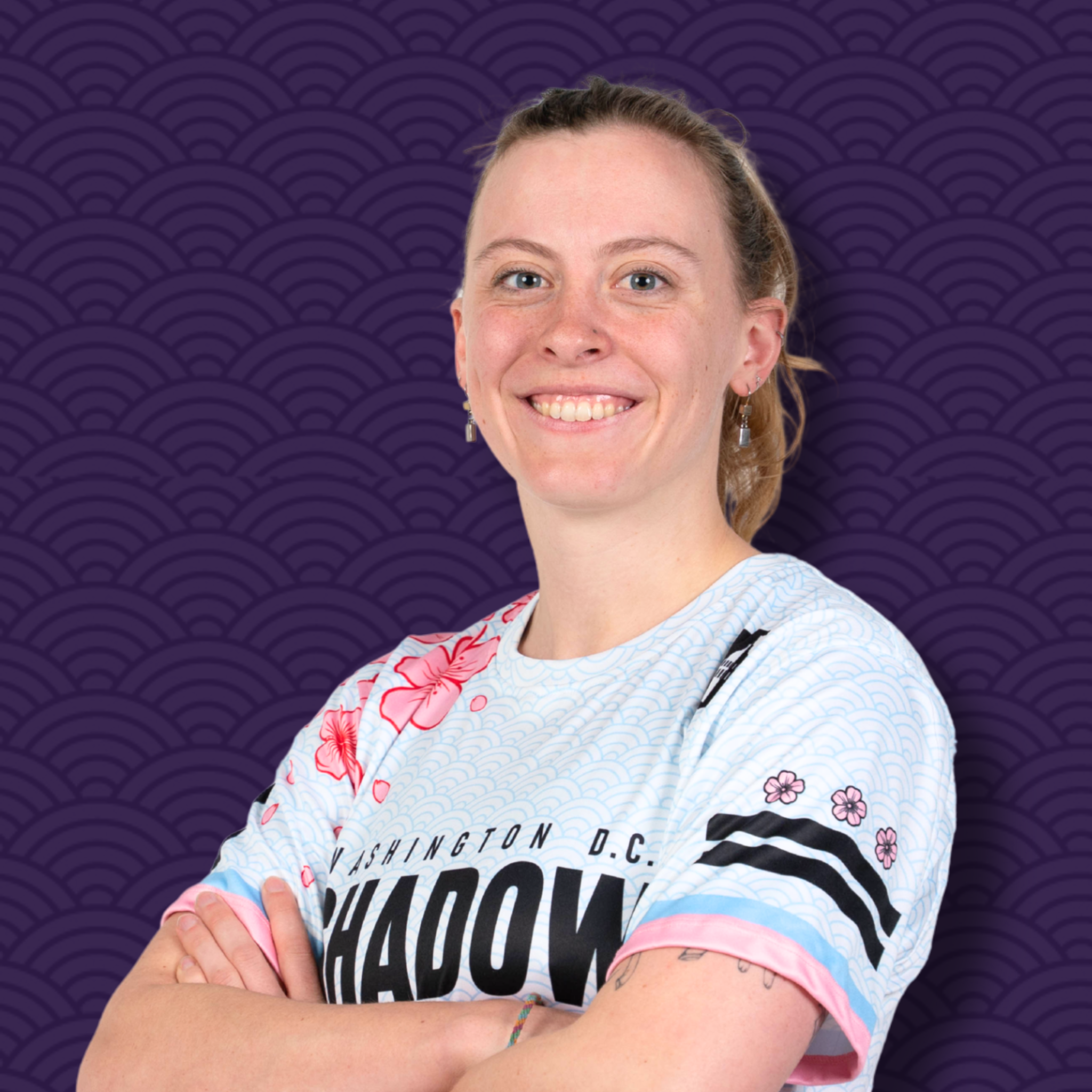 A woman with light skin and blue eyes, smiling, wearing a white t-shirt with black text, colorful floral pattern on the sleeves, and earrings, standing in front of a purple background with an Asian wave pattern.