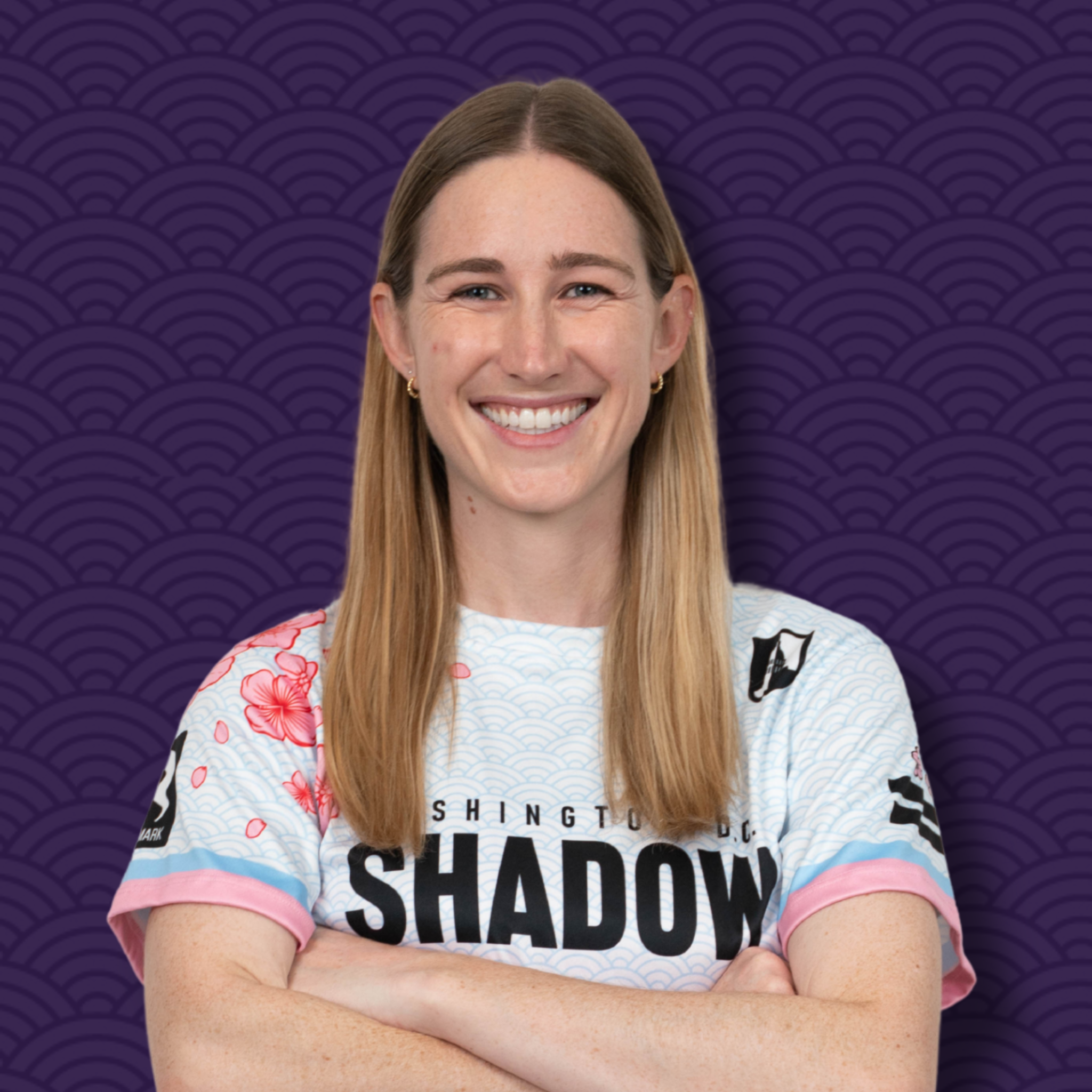 A young woman with long blonde hair smiling with crossed arms, wearing a white sports jersey with pink and black accents and the words 'SHANGTING SHADOW' on it, against a purple patterned background.