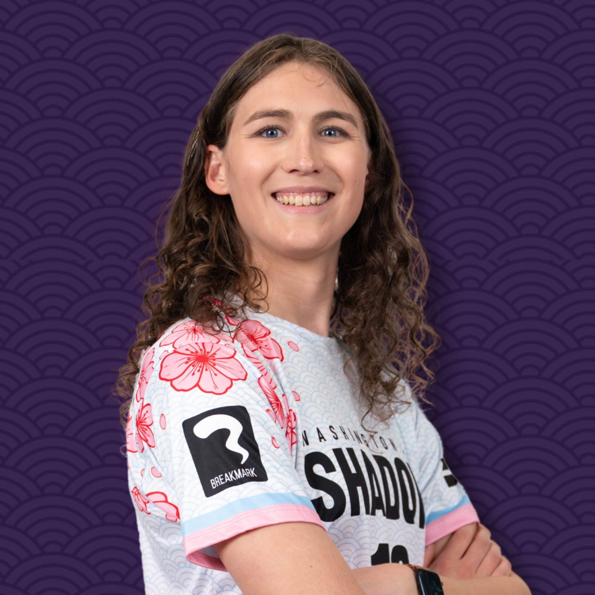 A young woman with curly brown hair and blue eyes smiling, wearing a white T-shirt with pink floral and black text, standing against a purple patterned background.