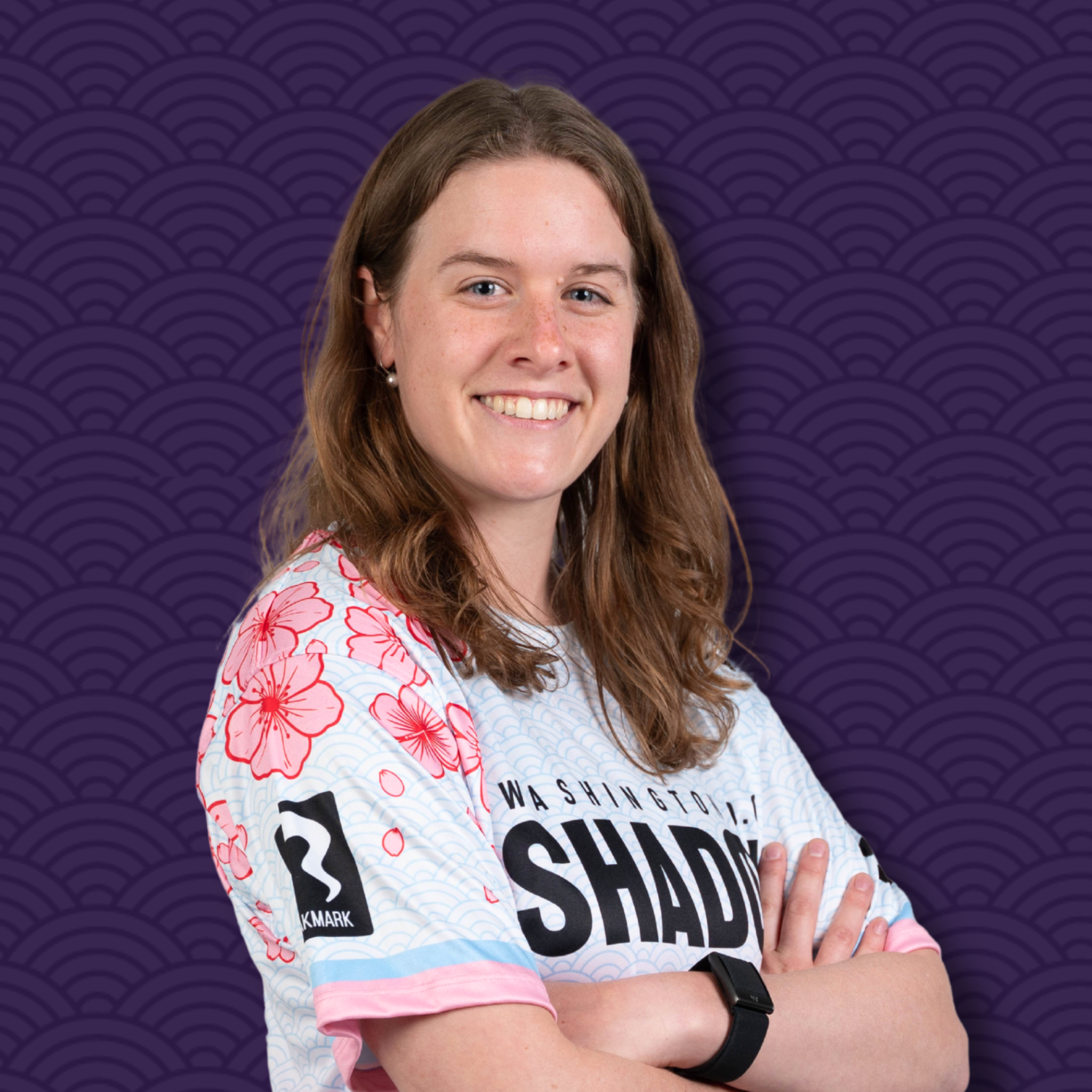 A woman with long brown hair smiling, wearing a white shirt with pink and red floral patterns, and a black smartwatch on her wrist, standing against a purple background with a geometric pattern.