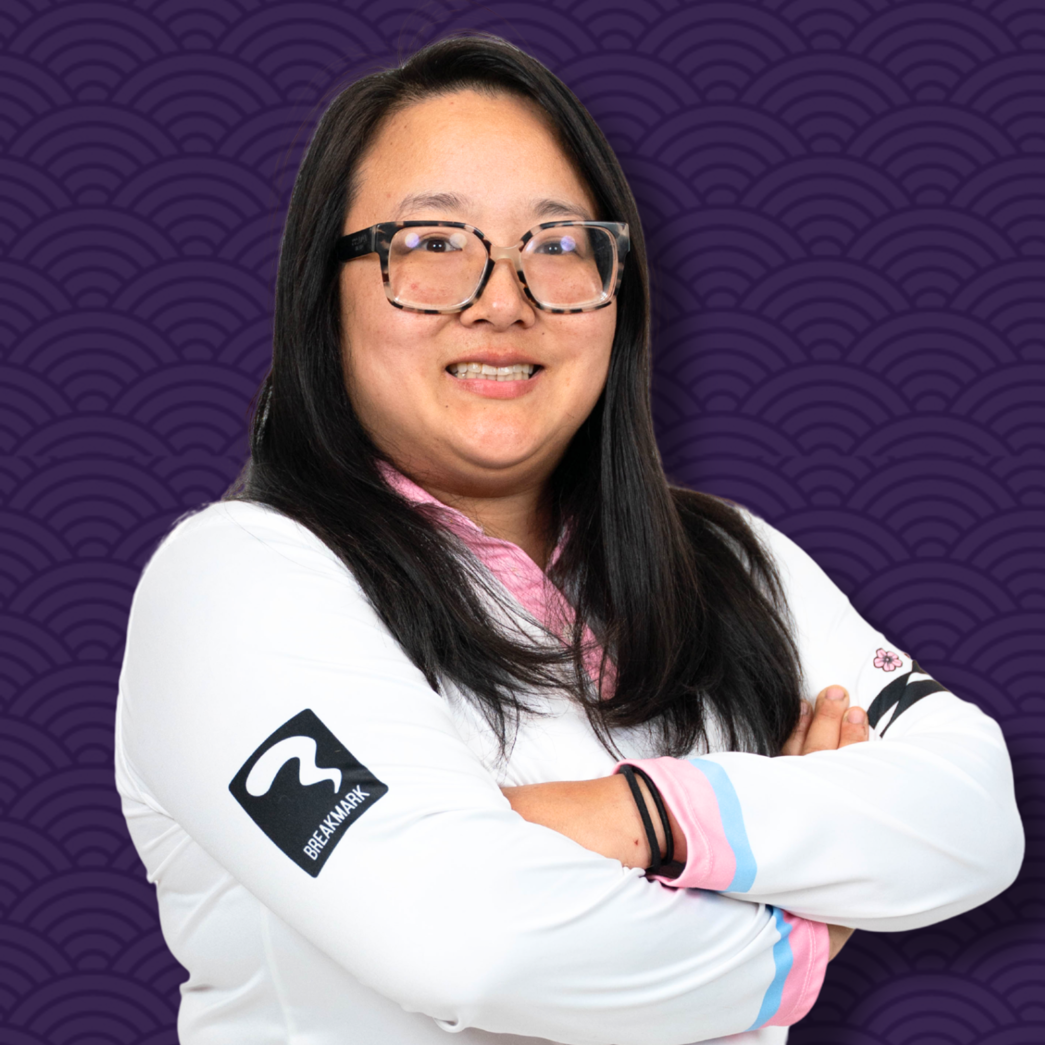 Close-up of a smiling young woman with long dark hair and glasses, crossing her arms, wearing a white jacket with black, pink, and blue accents, and a black wristband, standing against a purple background with a wave pattern.