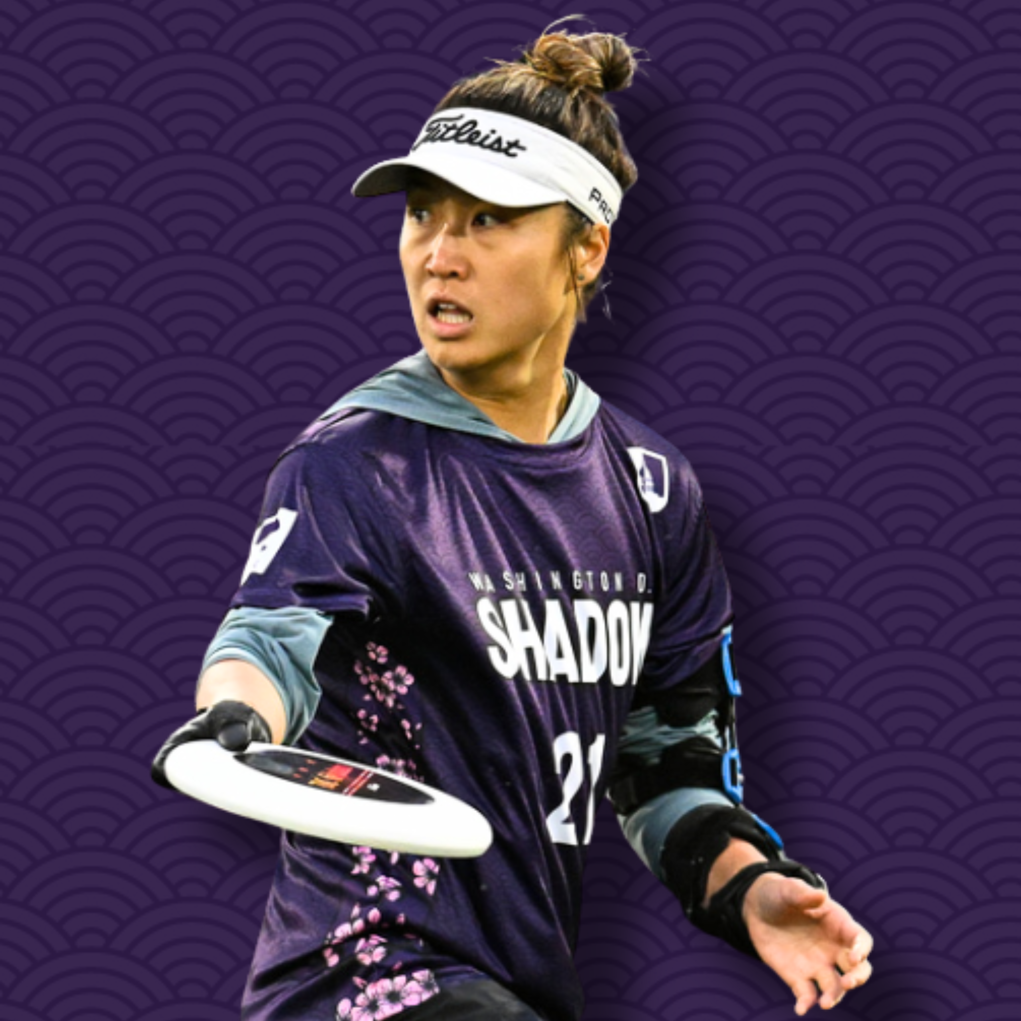 A person wearing a purple jacket, a floral shirt, black gloves, a white visor with the brand 'Titleist', and holding a golf club, standing against a purple patterned background.