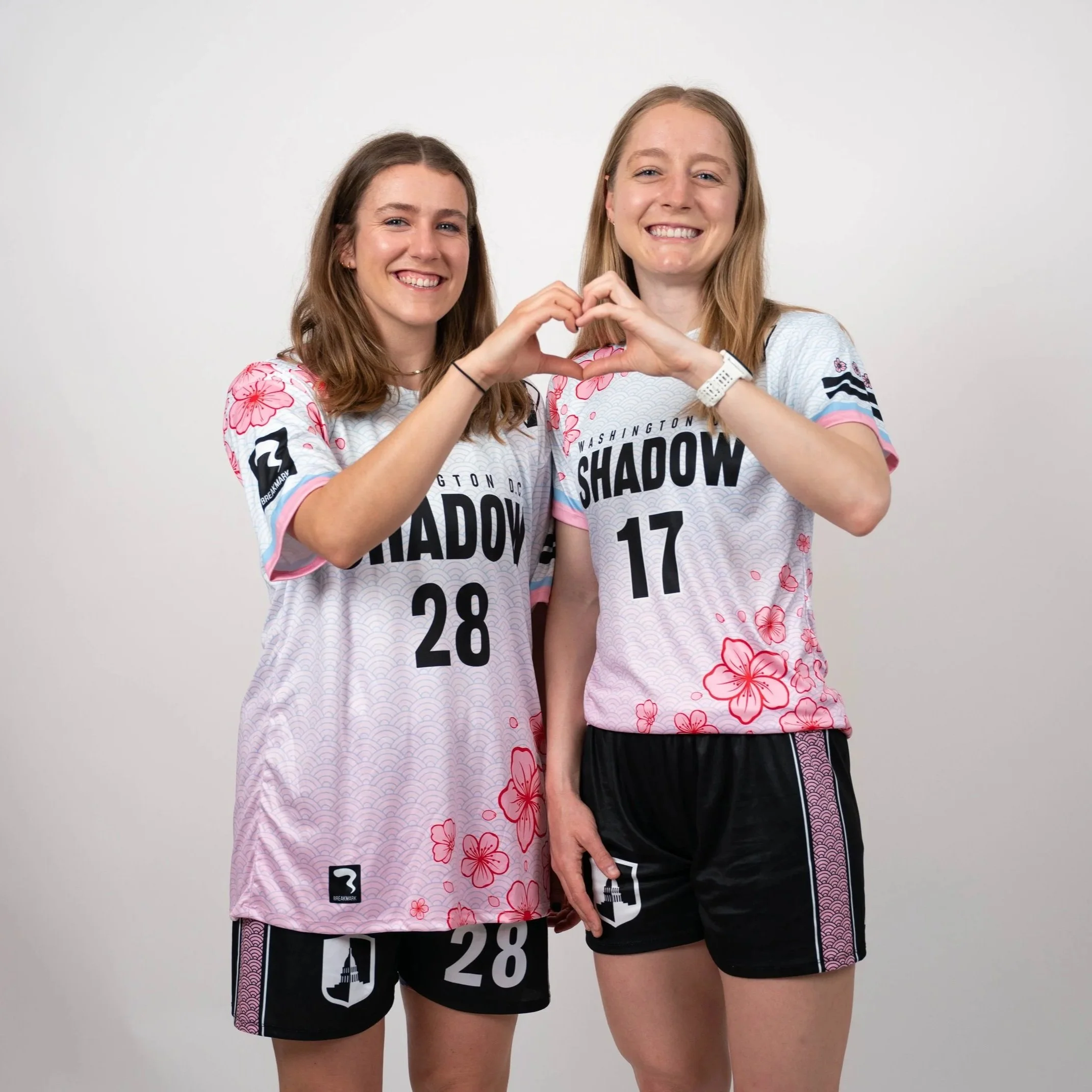 Two smiling women wearing matching sports jerseys, making a heart shape with their hands, standing against a plain white background.
