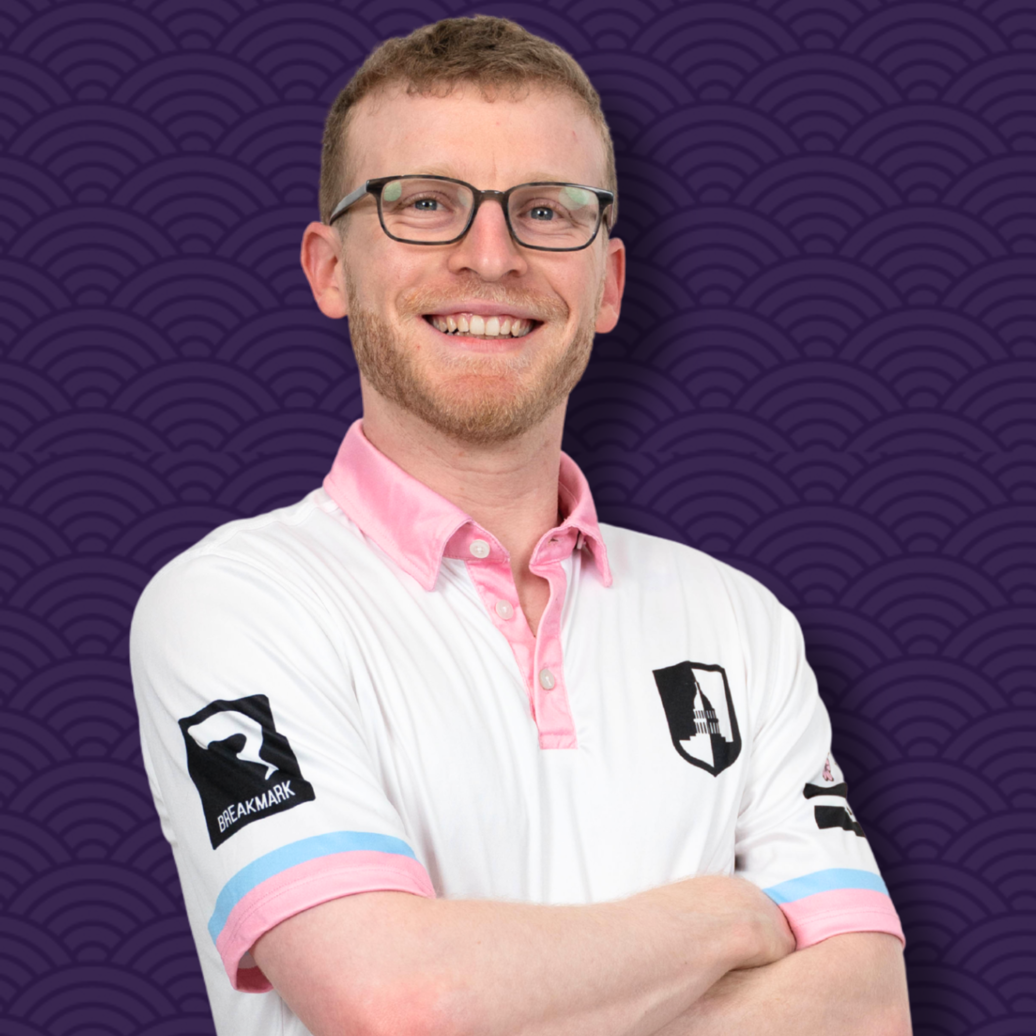 A man with short light brown hair, glasses, and a light beard smiling with crossed arms, wearing a white jacket with black text on the sleeves and a pink collar, against a purple background with a repeating wave pattern.