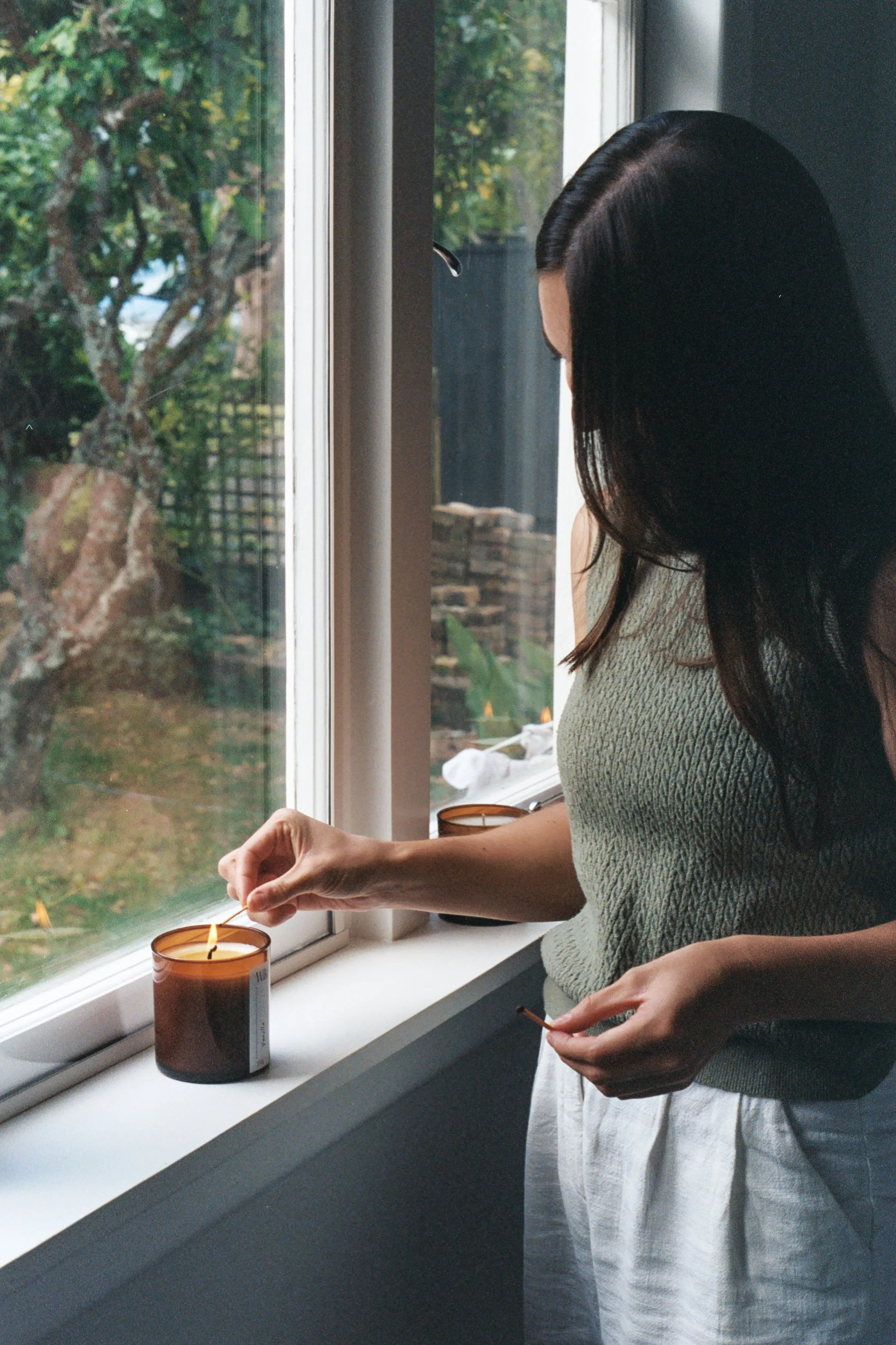 A woman with long dark hair, is lighting a Wik candle by a window. Outside, trees are visible. The woman is holding a matchstick, and the candle is in an amber glass jar on the windowsill.