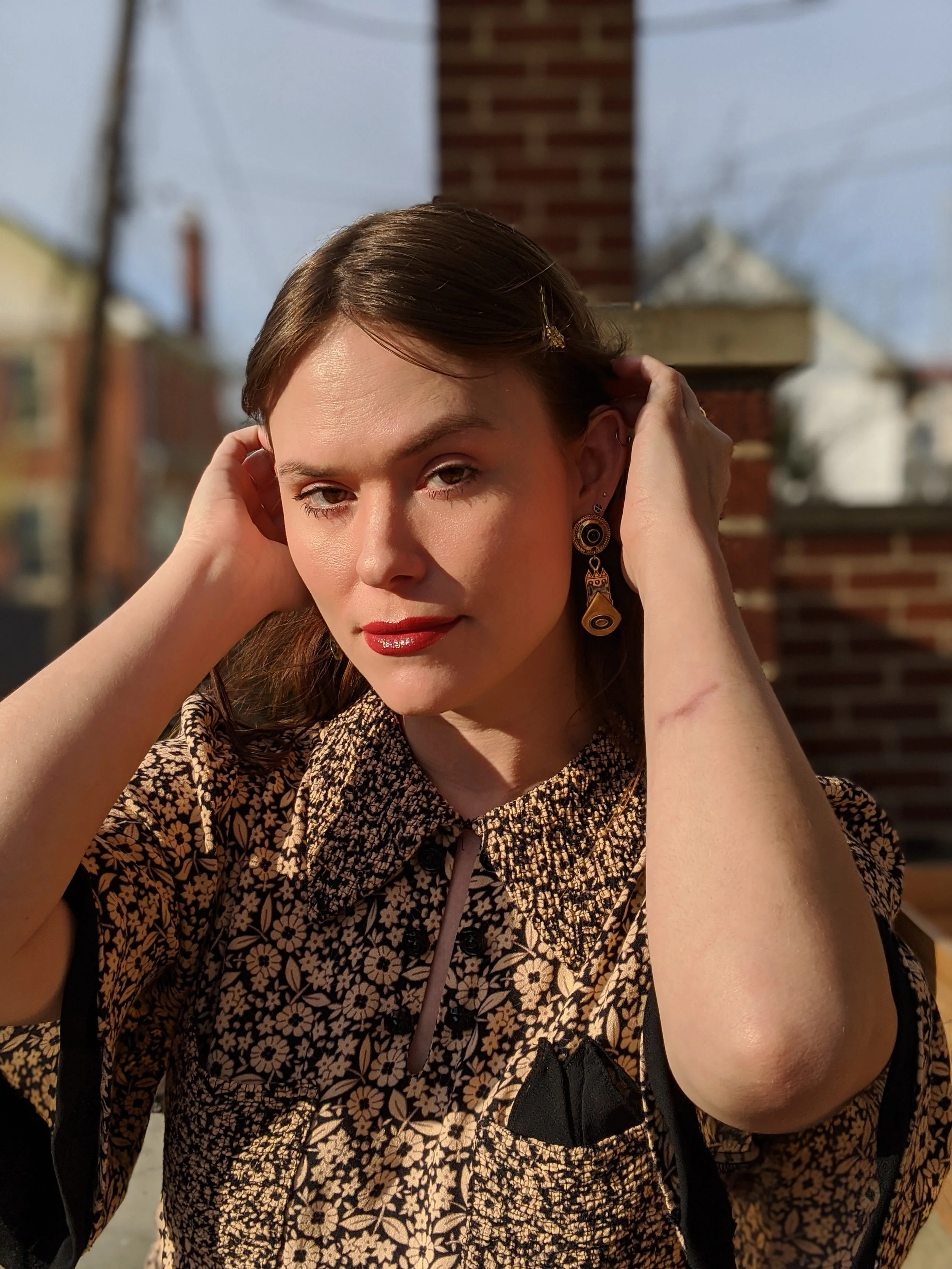 Close-up of a woman with brown hair, wearing earrings, a patterned blouse, adjusting her hair with both hands, outdoors with a brick wall in the background.