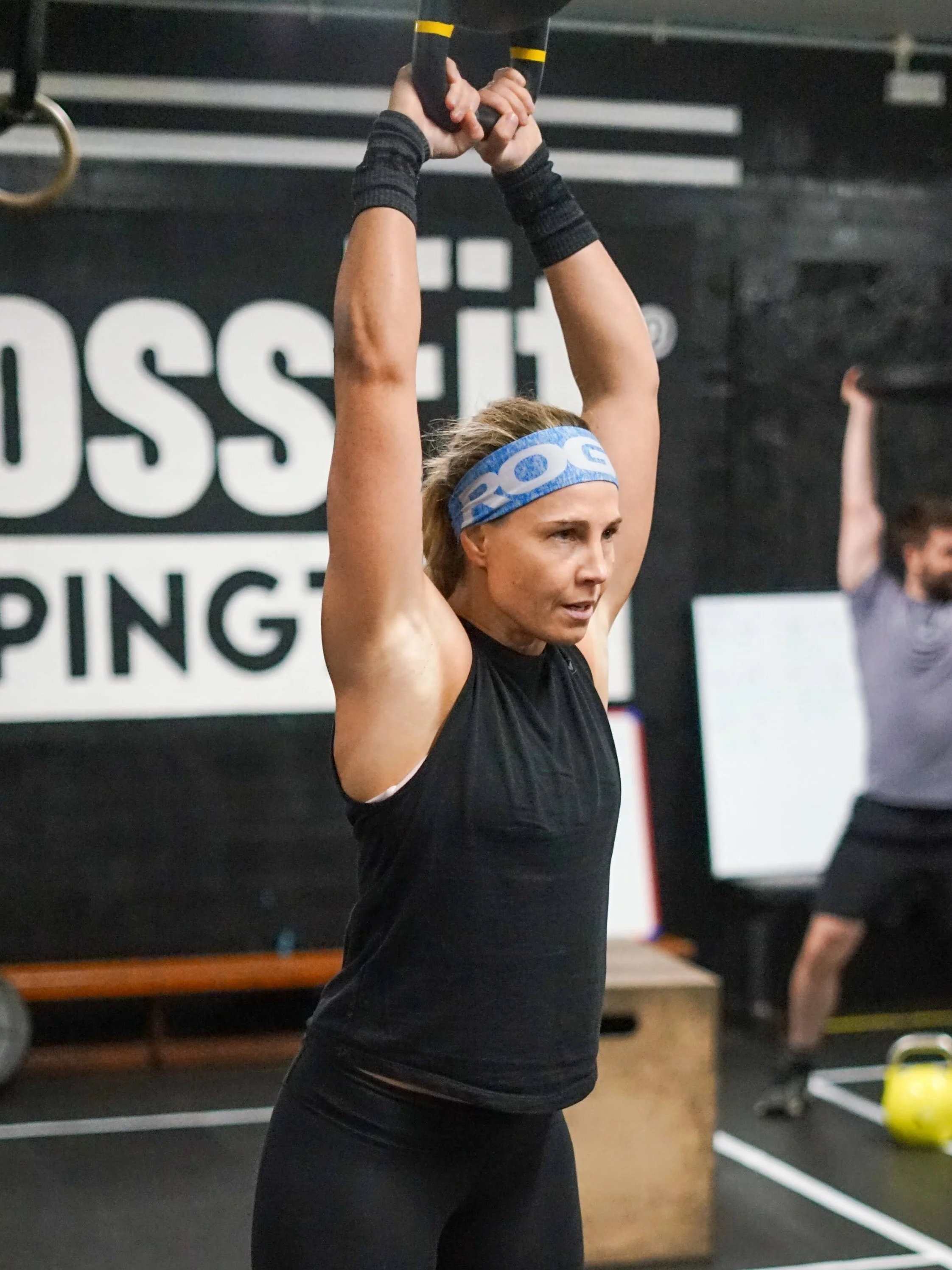 A woman in a black tank top and leggings, wearing a blue Reebok headband, is lifting an exercise bar above her head in a gym.
