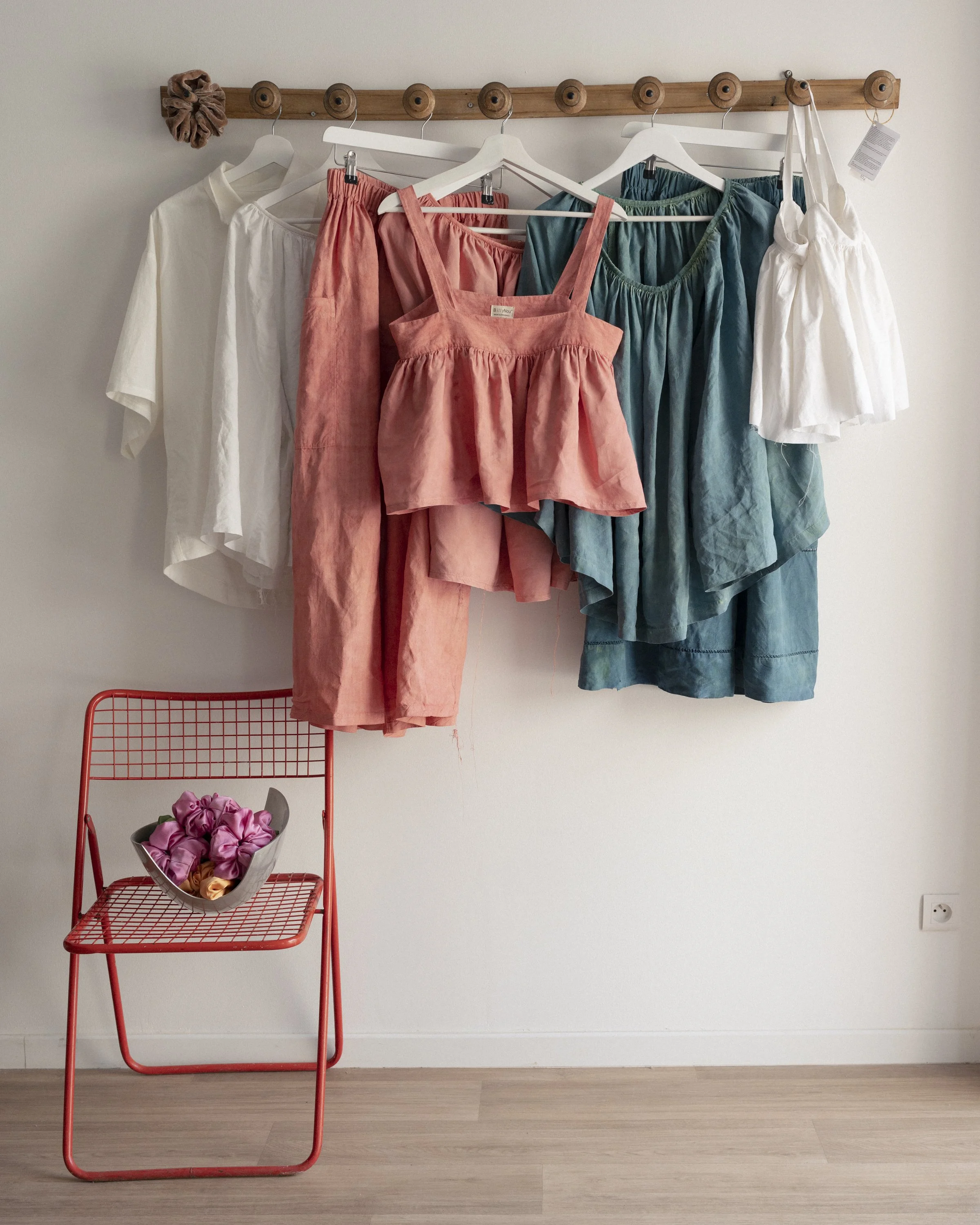 Clothes hanging on a wooden coat rack, including white, pink, teal, and green garments, next to a red wire chair with a bowl of pink and purple fabric scraps.