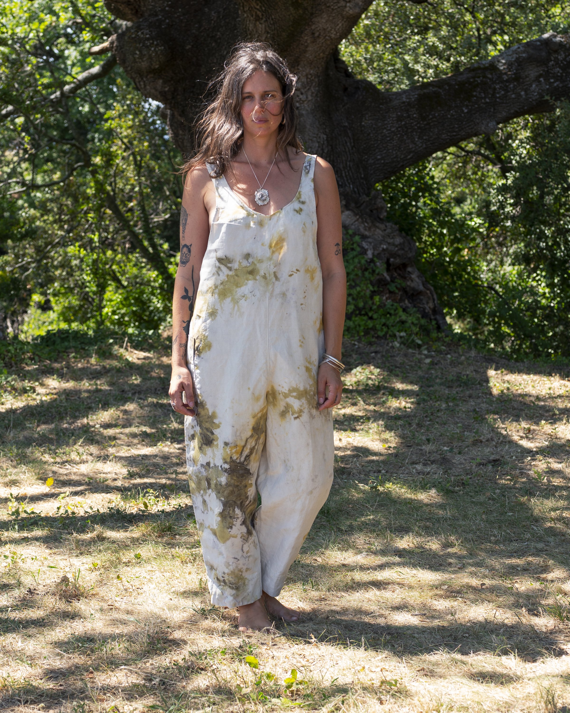 A woman standing barefoot on grass in front of a large tree with green foliage, wearing a white and yellow tie-dye jumpsuit with tattoos on her arms and accessorized with jewelry.
