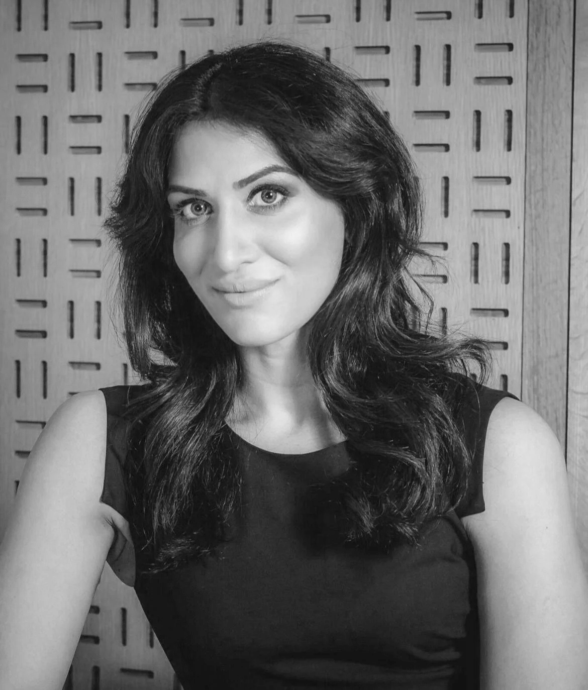 Black and white portrait of a woman with wavy hair, wearing a sleeveless top, smiling subtly in front of a patterned wooden background.