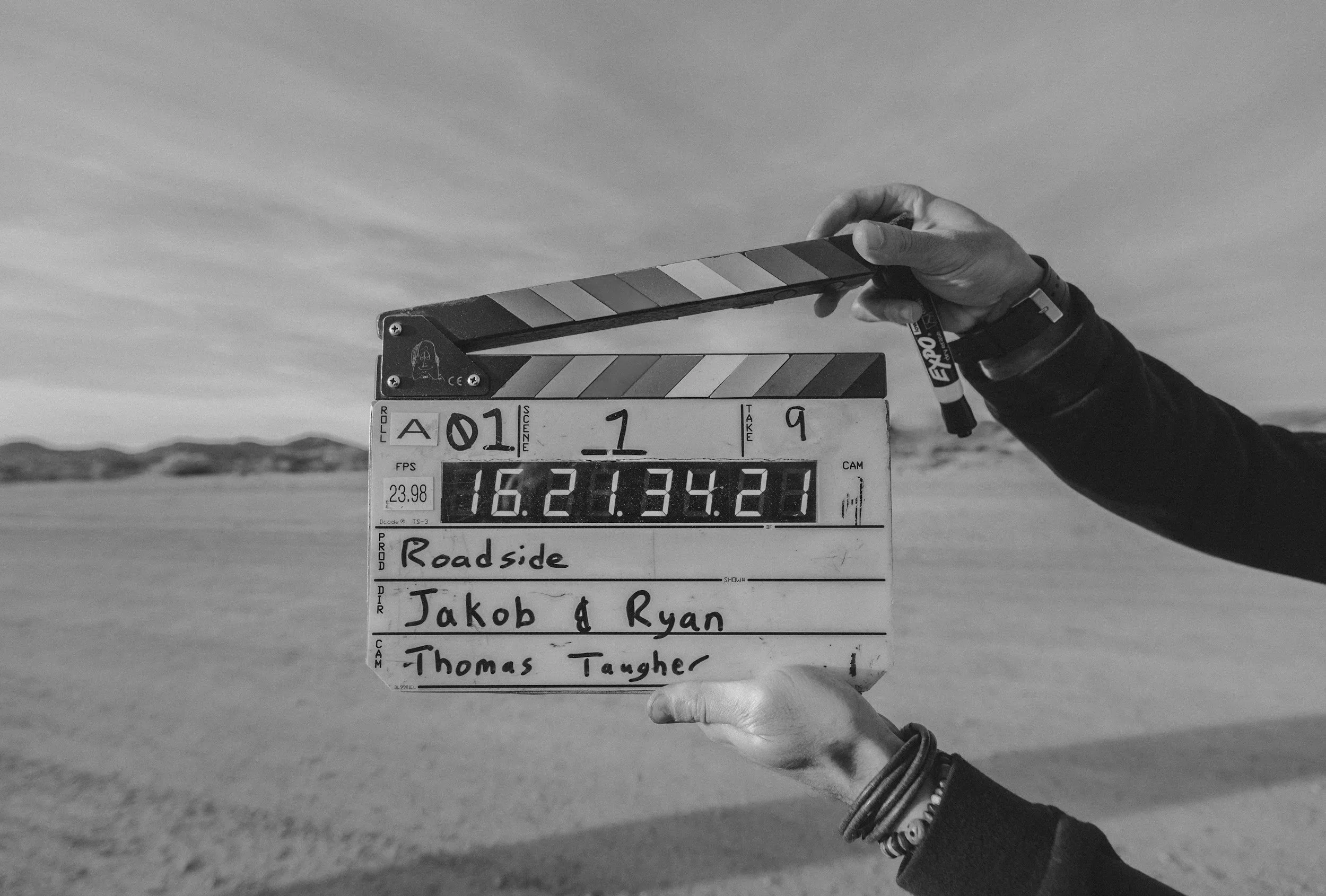 Black and white photo of a person's hand holding a film slate, with a landscape in the background. The slate has text including titles and names, indicating the start of filming for a project.