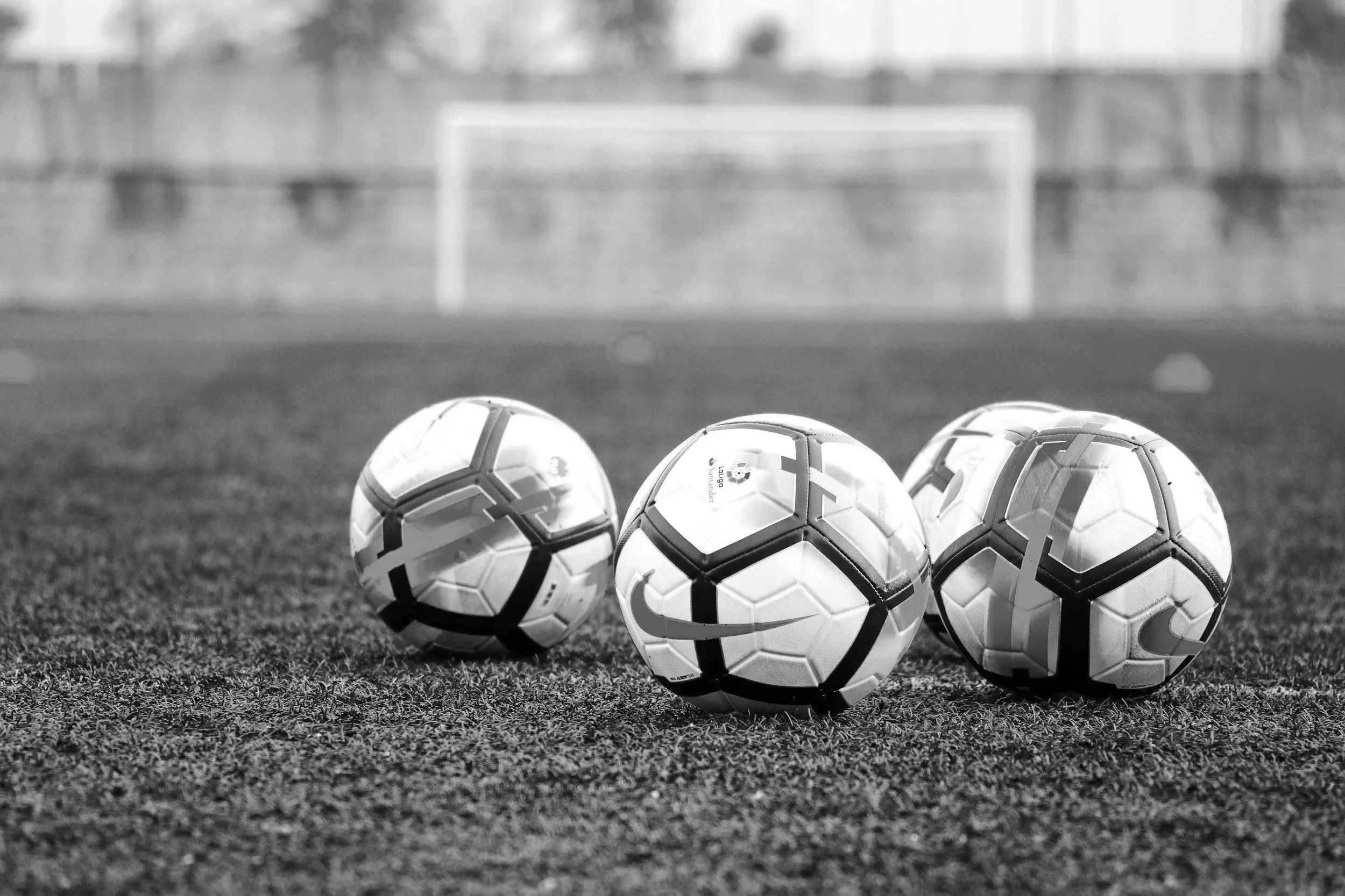 Four soccer balls are placed on a grass field with goals and trees visible in the background. The image is in black and white.