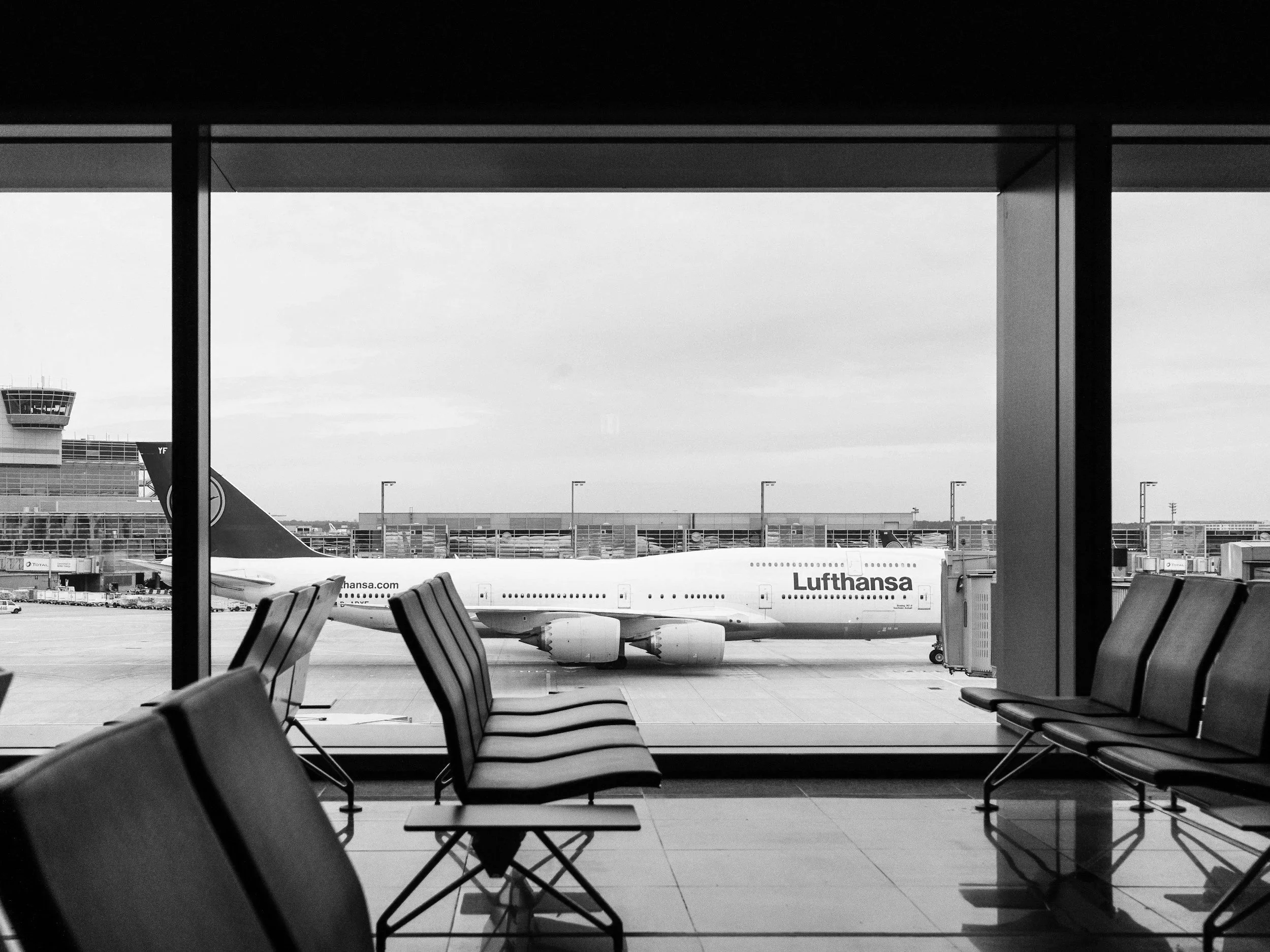 Empty airport terminal seating area with chairs facing a large window. Outside the window, an airplane with Lufthansa branding is parked at the gate, and airport buildings and control tower are visible in the background.